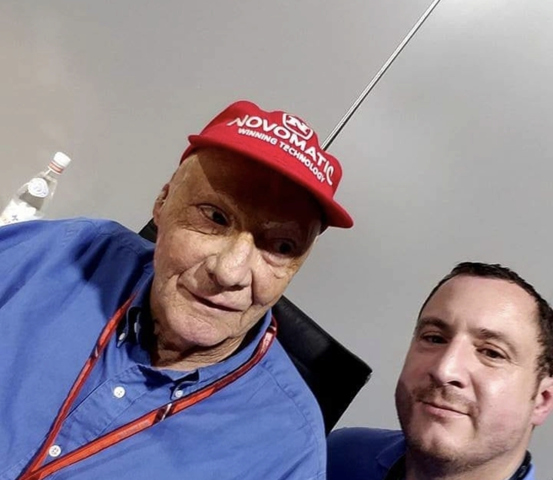Two men, one older with a red cap and blue shirt, and one younger, smiling, in an indoor setting.