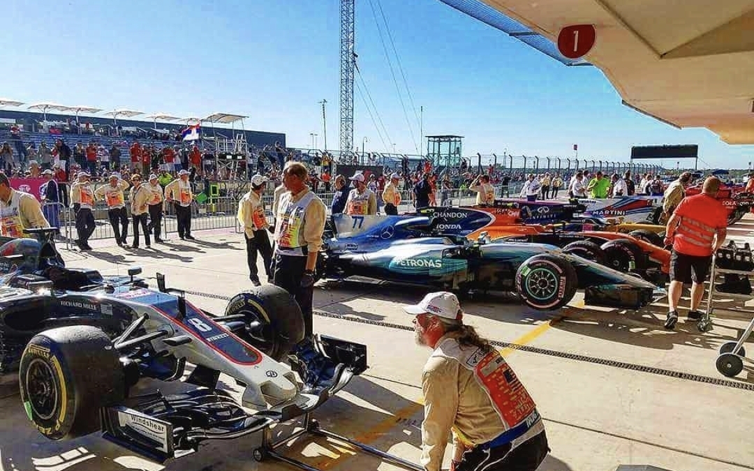 Formula 1 cars in pit lane with team members and spectators in background under a clear sky.