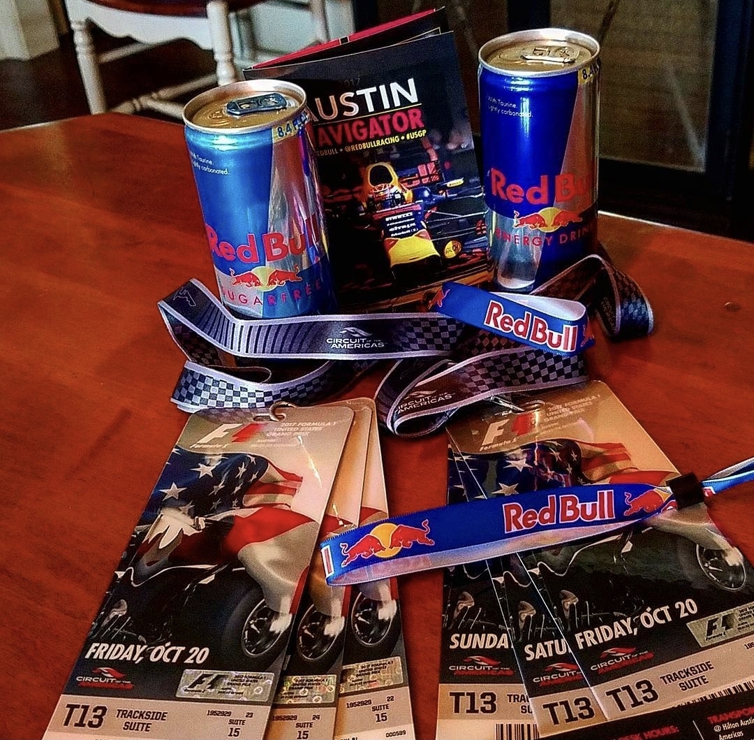 Two Red Bull energy drink cans, race tickets, and a lanyard on a wooden table, with a magazine in the background.
