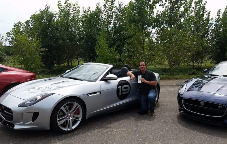 Two men kneel beside a silver sports car with a black circle and number 19 on the door, parked outdoors with trees in the background.