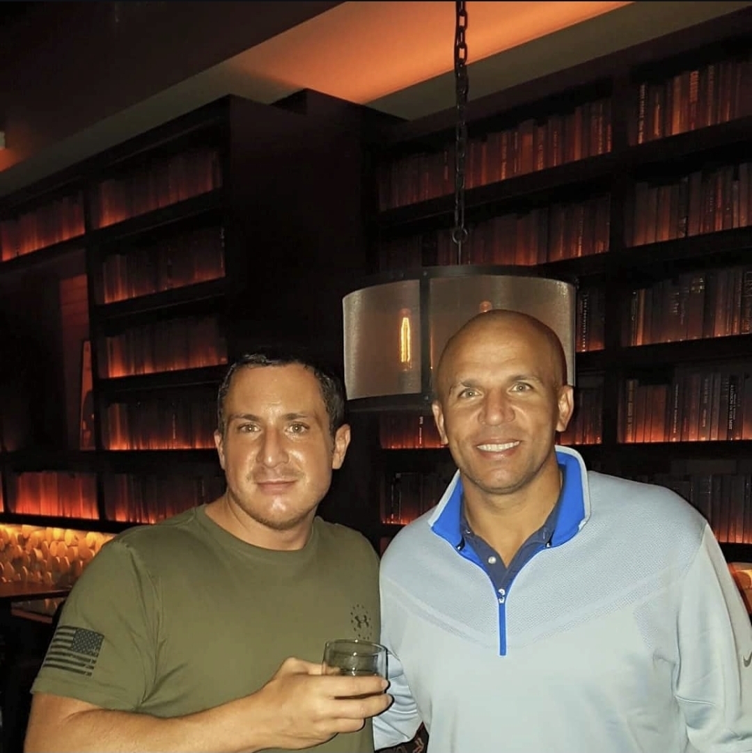 Two men standing indoors, one holding a glass, in front of a dark bookshelf with warm lighting.