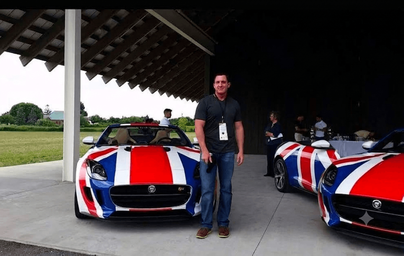 Man standing next to a sports car with red, white, and blue stripes, under a large dark structure, with other cars nearby.