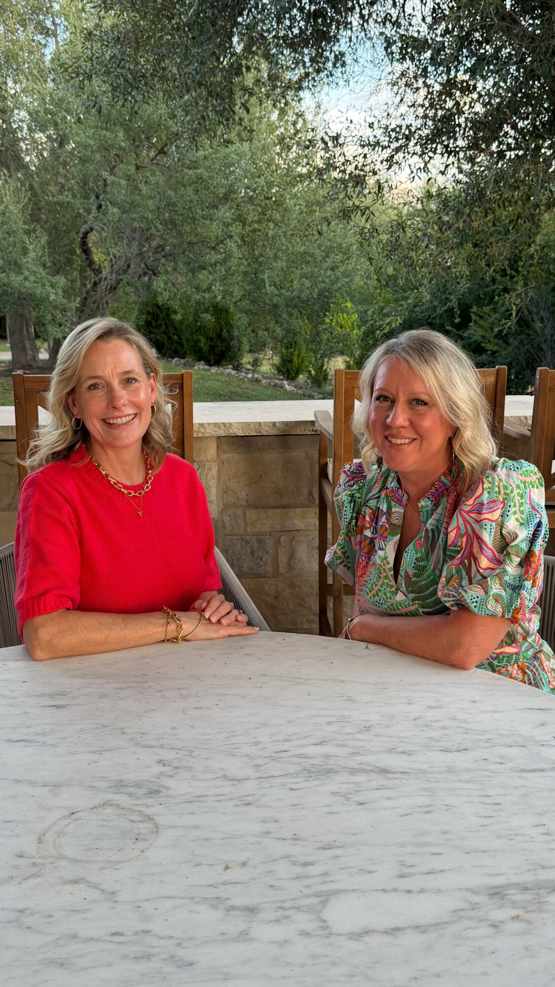 Two women sit at a table outdoors with trees in the background, smiling and facing the camera.
