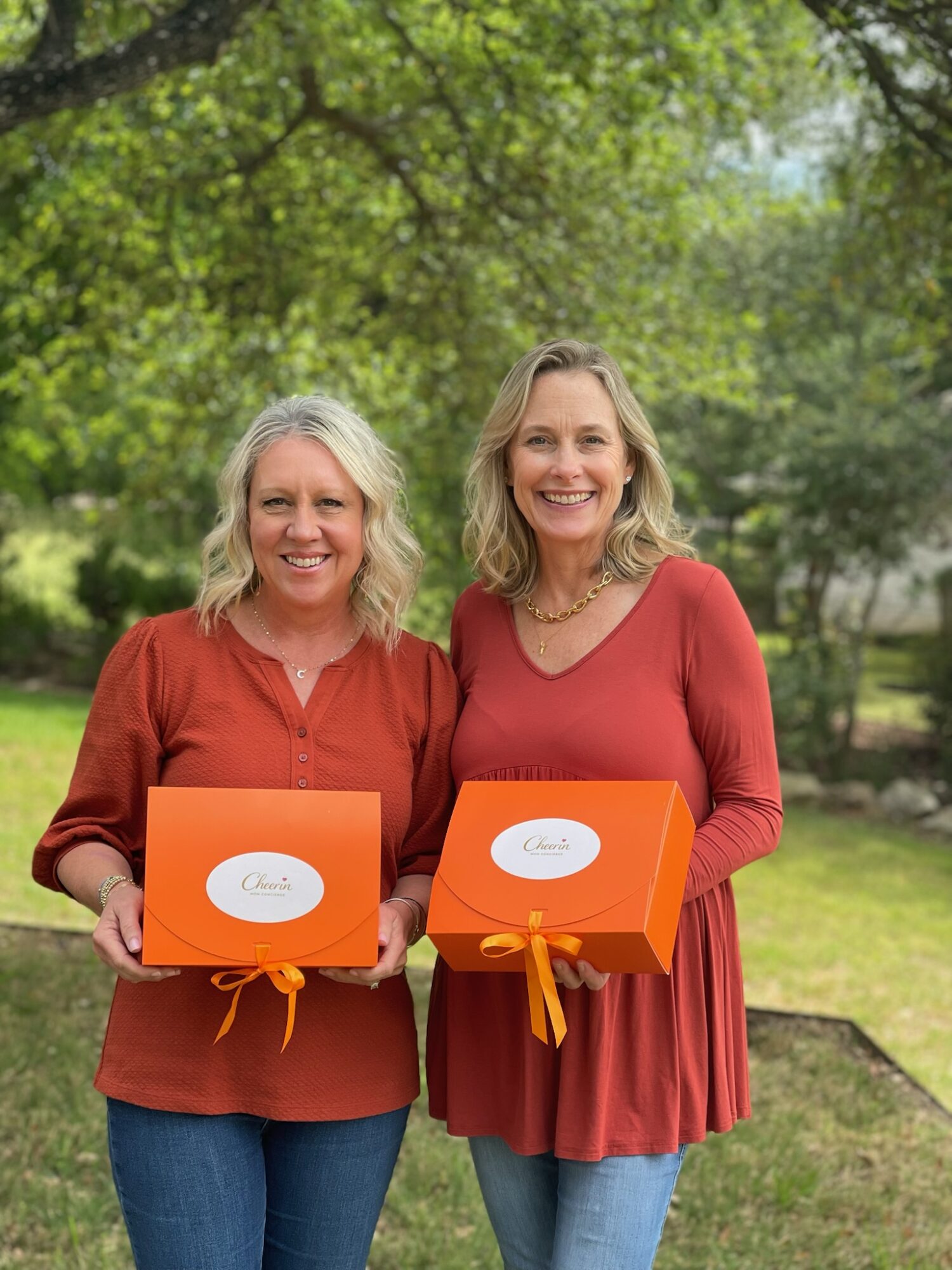 Two women smiling outdoors holding orange gift boxes with white labels, surrounded by green trees and grass.