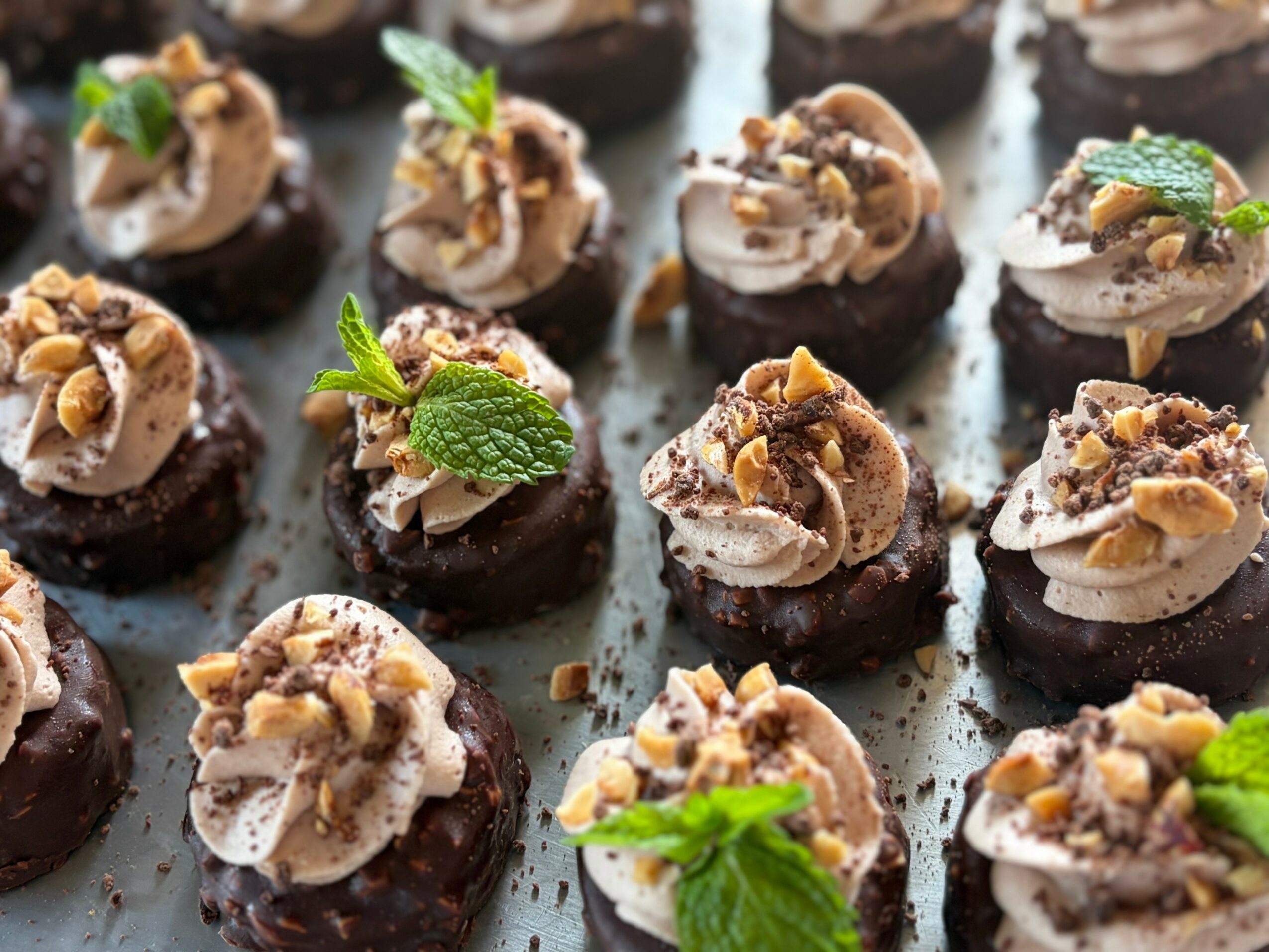 Small chocolate desserts topped with cream, nuts, and mint leaves, arranged on a tray.