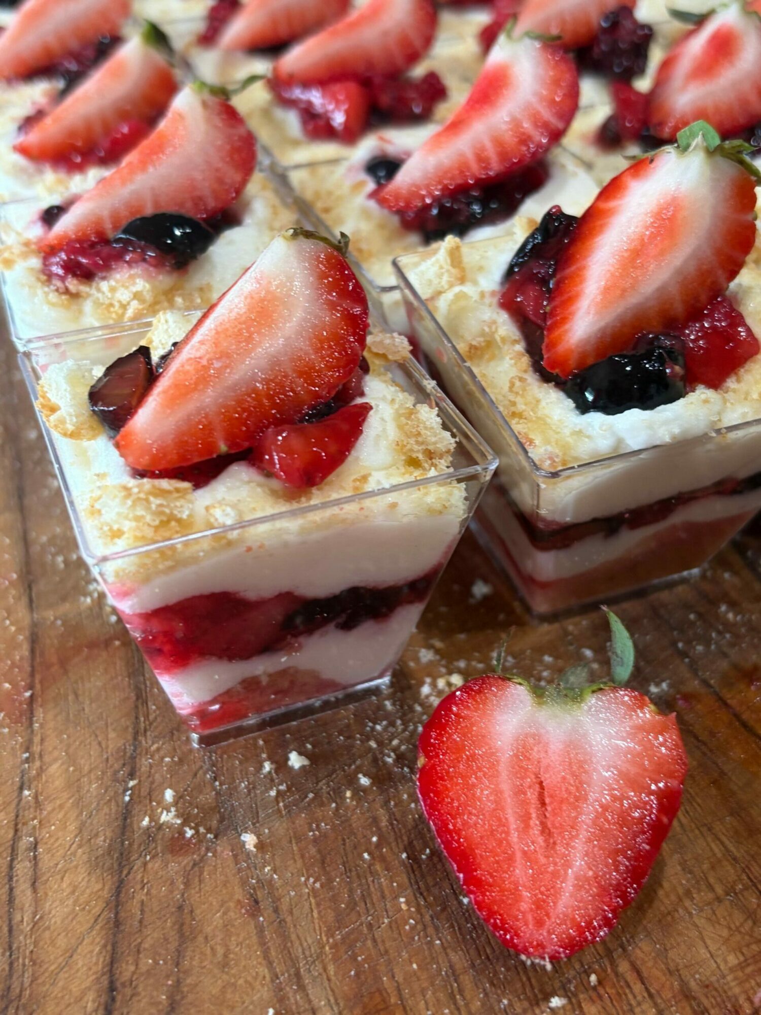 Slices of layered cake with strawberries and berries on top, on a wooden surface with a halved strawberry nearby.