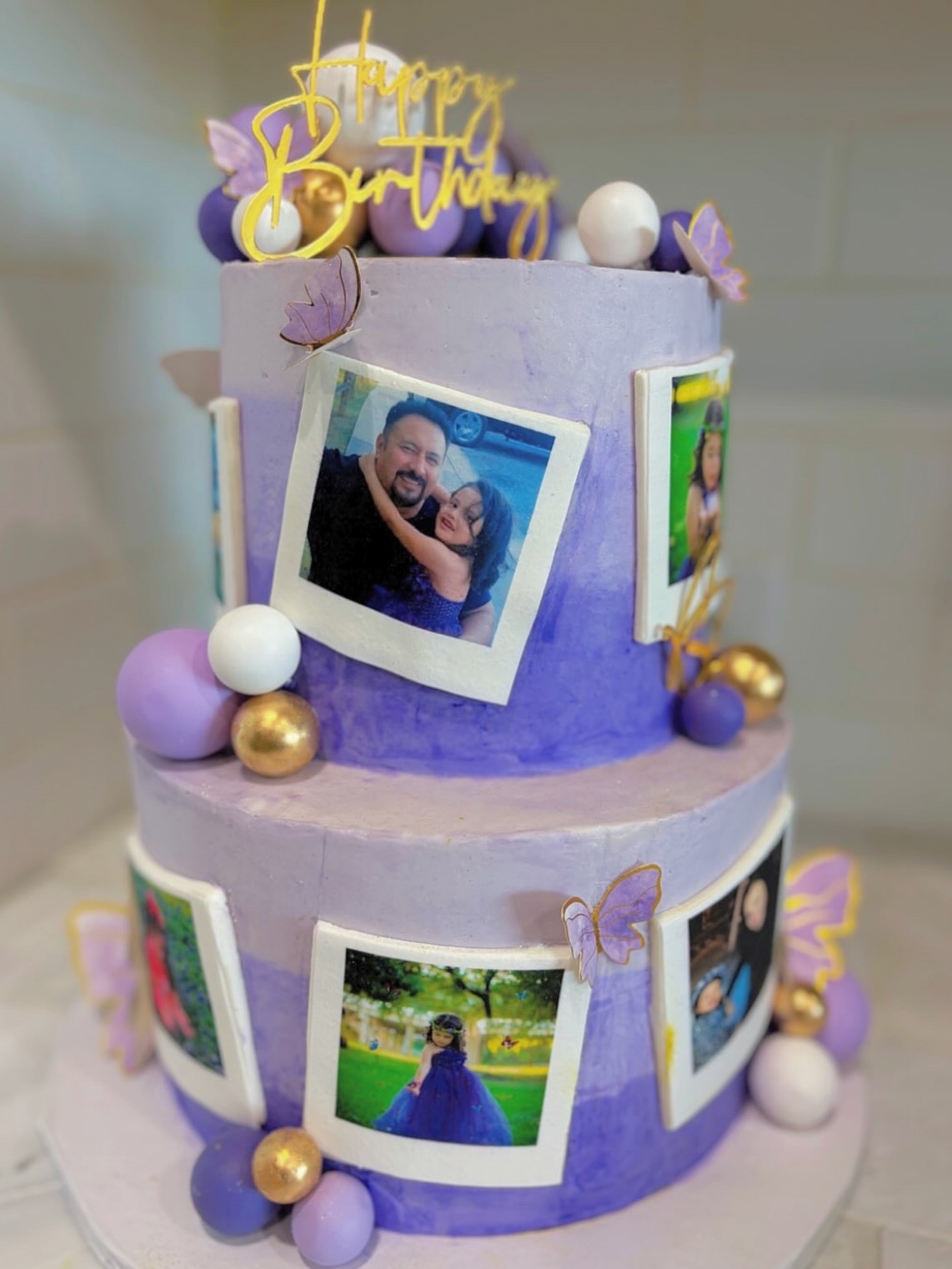 Two-tier purple birthday cake decorated with photos, purple and white balls, and a gold 'Happy Birthday' topper.