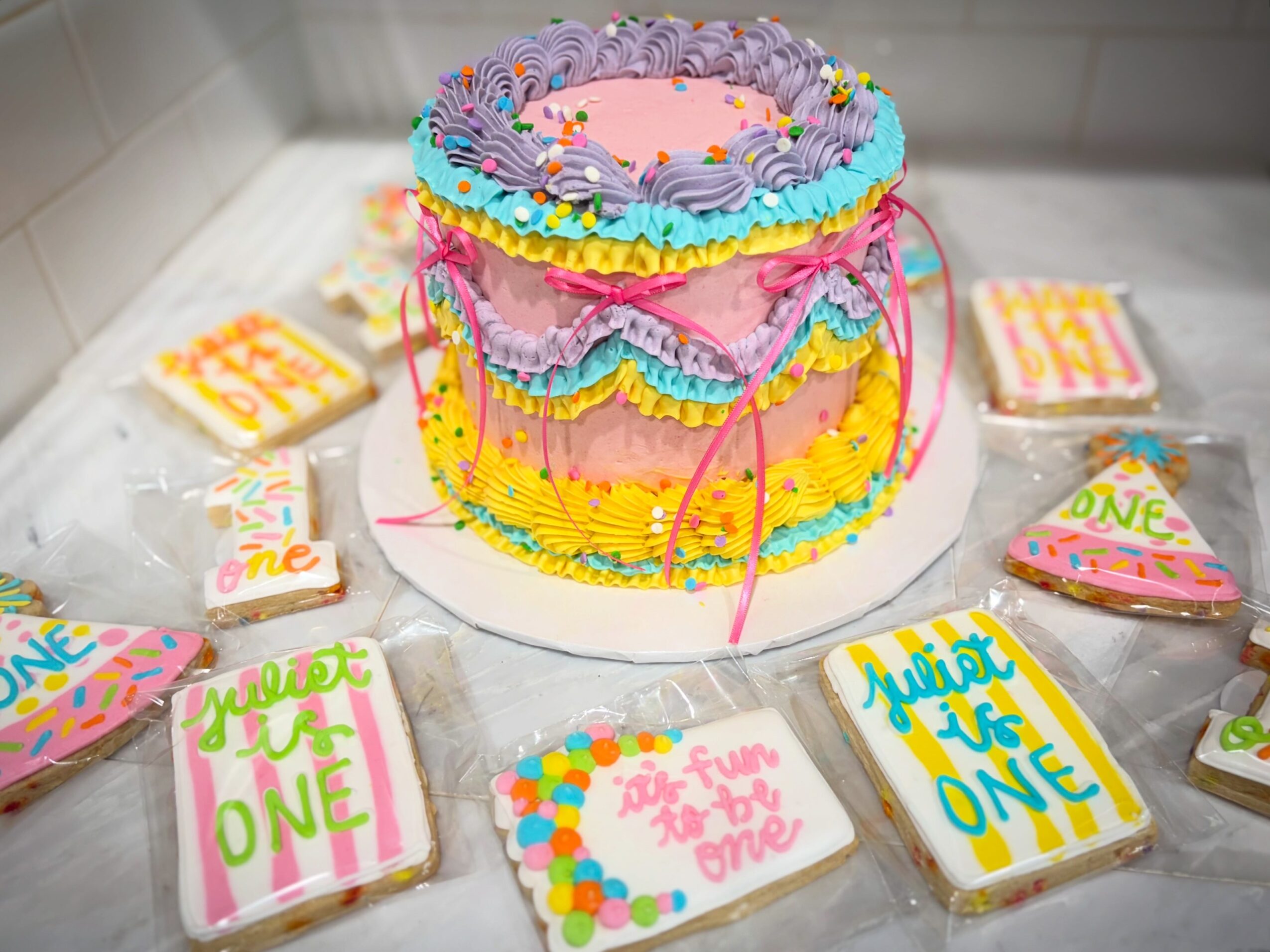 Colorful birthday cake with pink, purple, yellow, and blue icing, surrounded by decorated cookies, on a white surface.