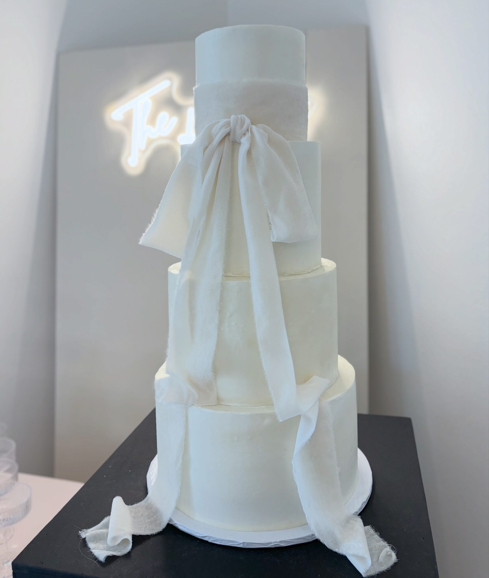 Three-tiered white cake with fabric draping, on a black table, with a neon sign in the background.