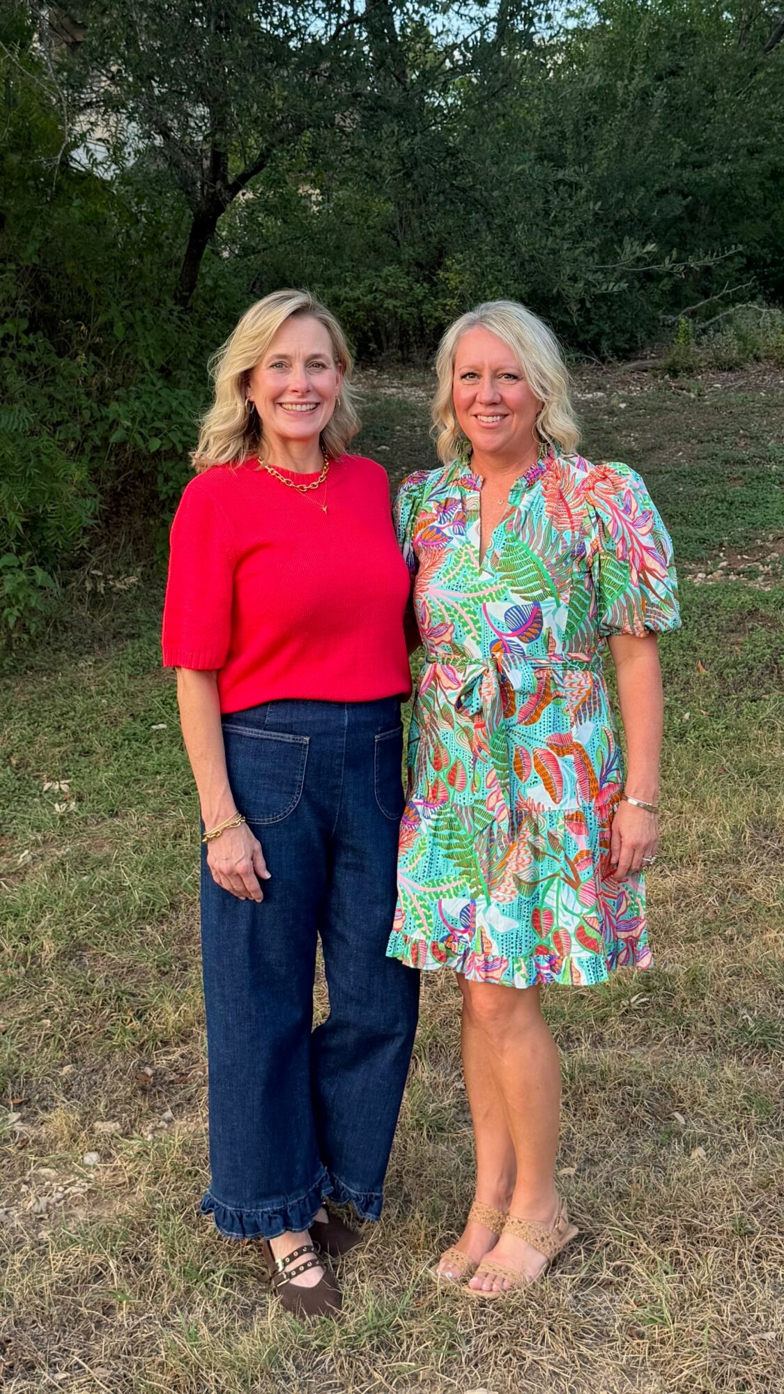 Two women stand outdoors on grass with trees in background, smiling, one in a red top and jeans, the other in a colorful dress.