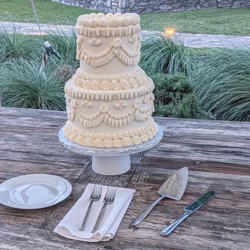 Three-tiered cake with decorative icing, placed on a cake stand outdoors, with a plate, fork, knife, and spatula on a wooden table.