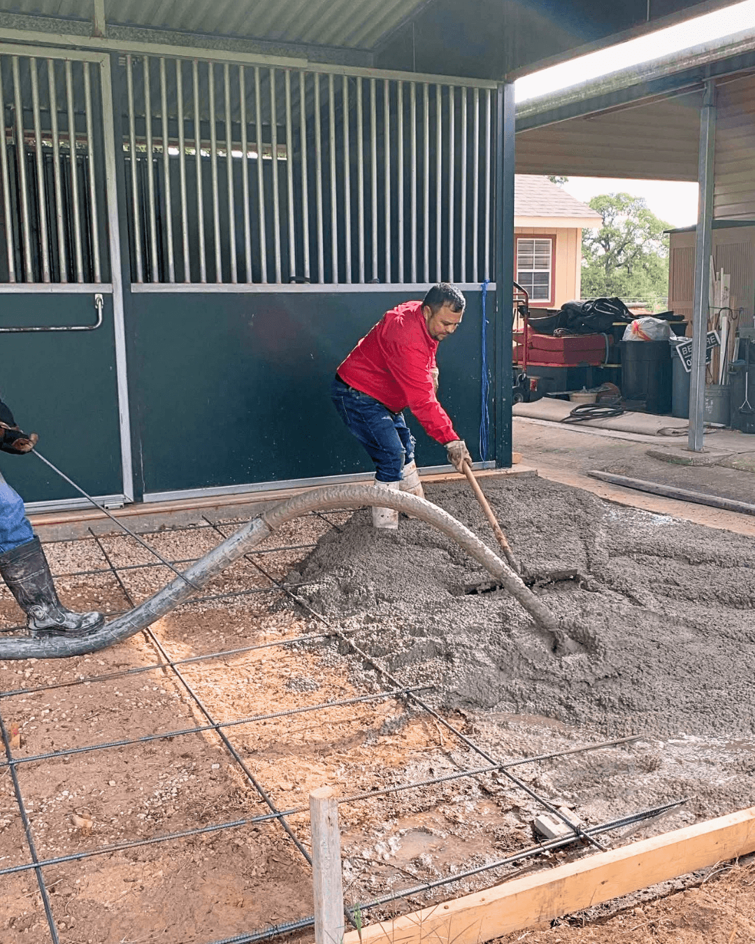 Person in red jacket spreading concrete with a tool on a construction site, with a building and parked vehicle in background.