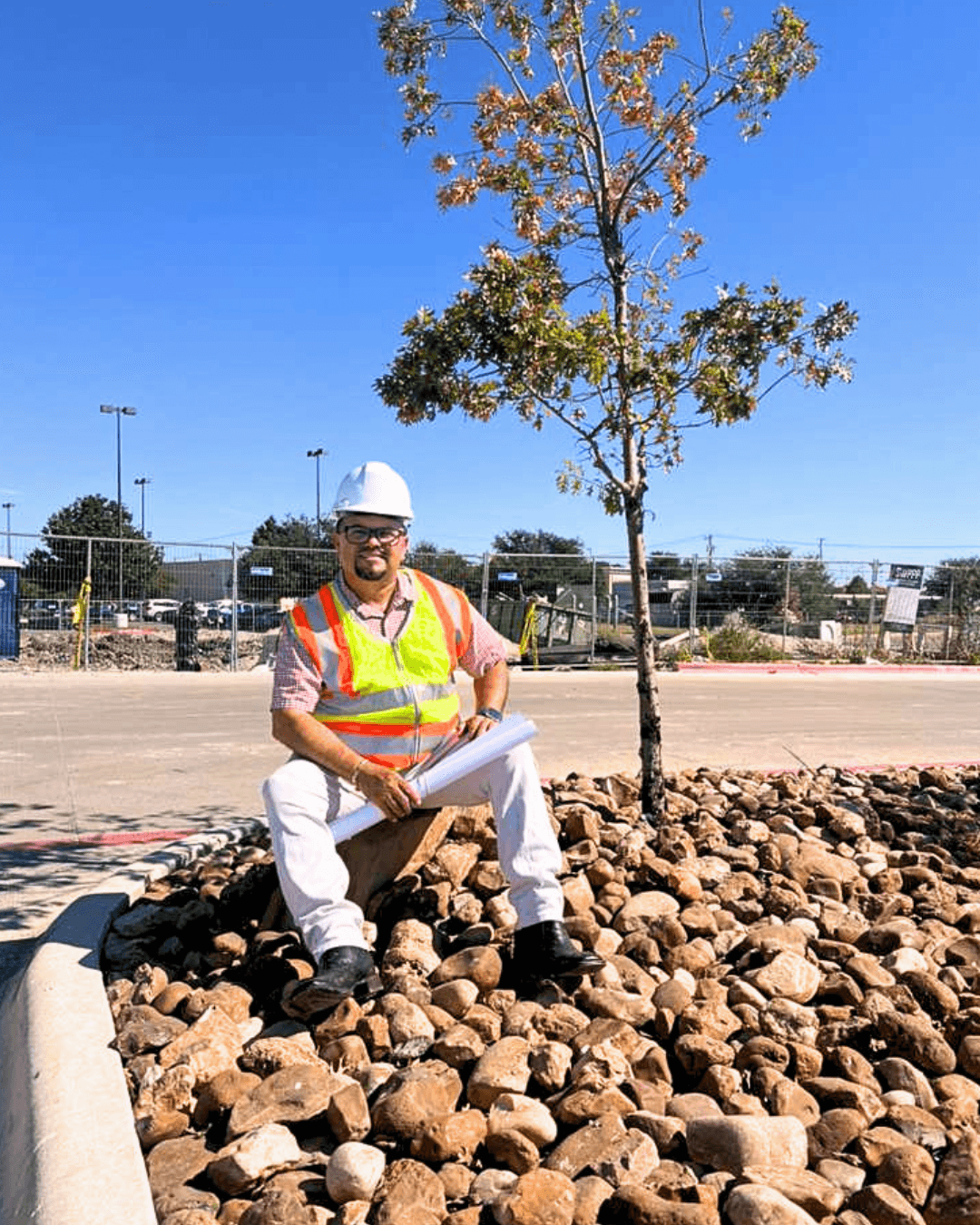 Construction worker in safety vest and helmet sitting on rocks near a small tree, with a fence and blue sky in background.