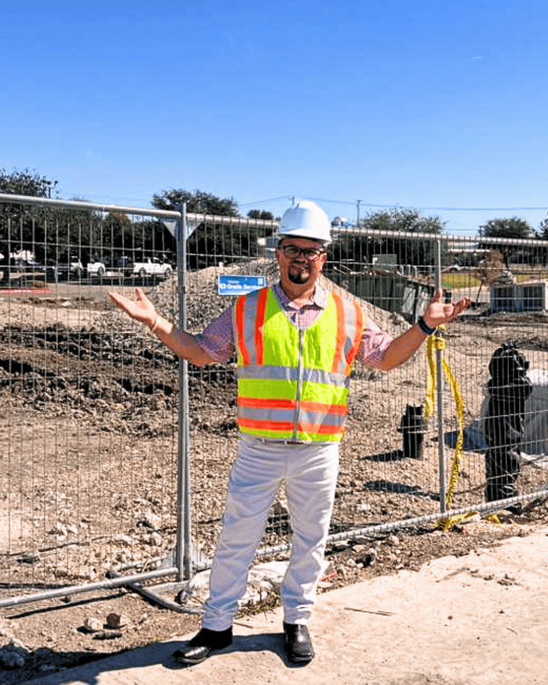 Person wearing a hard hat and safety vest standing outdoors with arms outstretched, construction site in background.