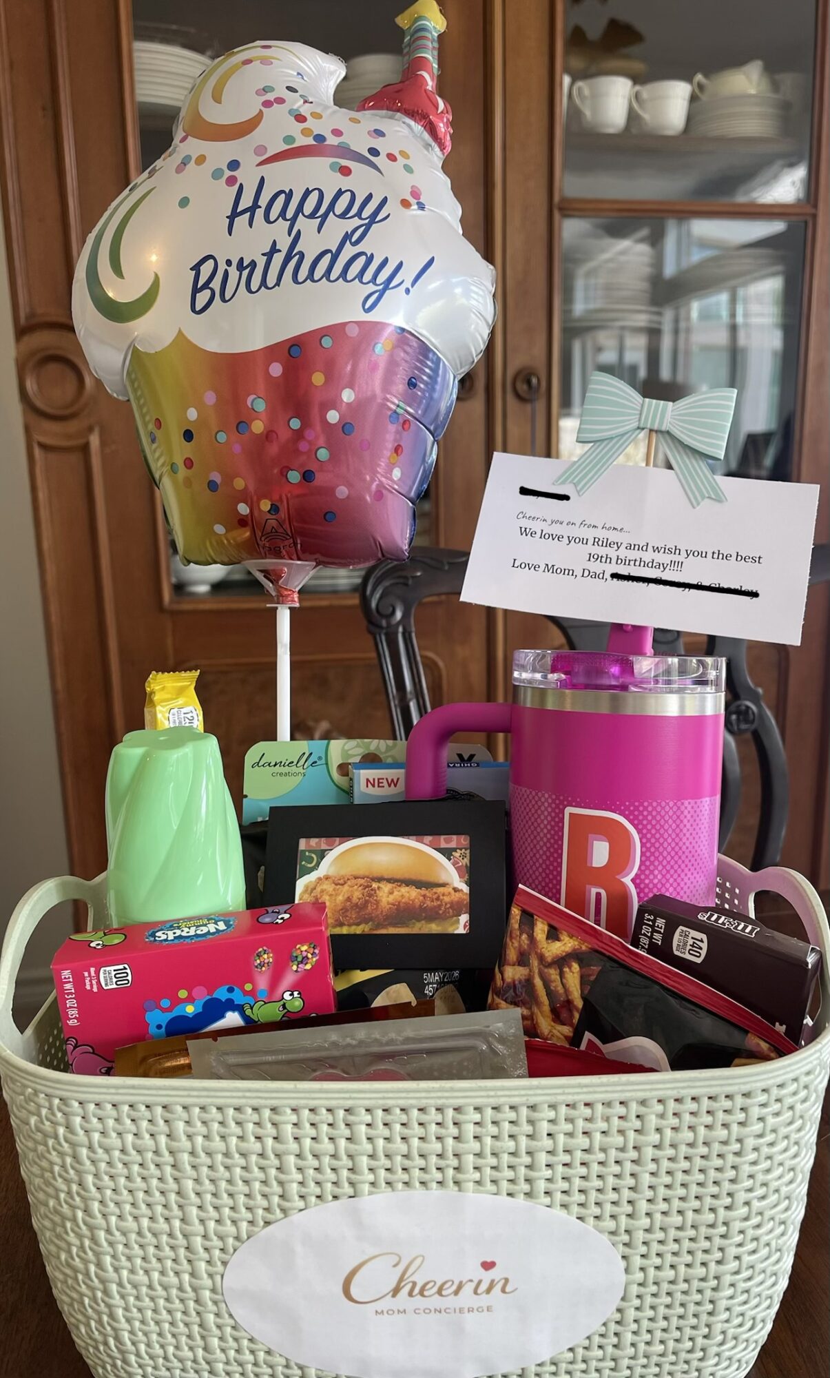 A birthday balloon, gift bag, and various items in a white basket on a wooden shelf.
