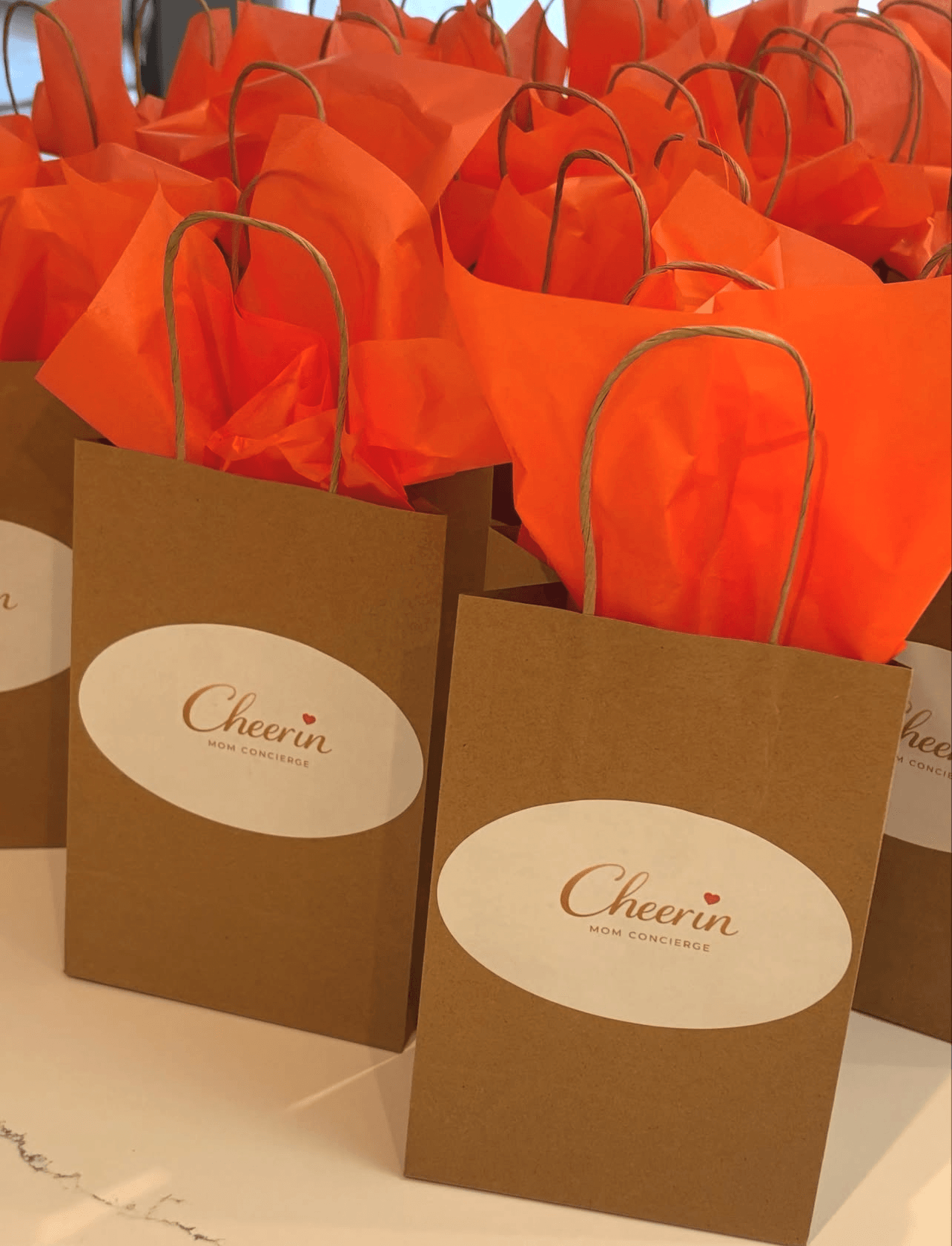 Brown paper gift bags with orange tissue paper and handles, labeled 'Cheerin' on white oval stickers, arranged on a table.