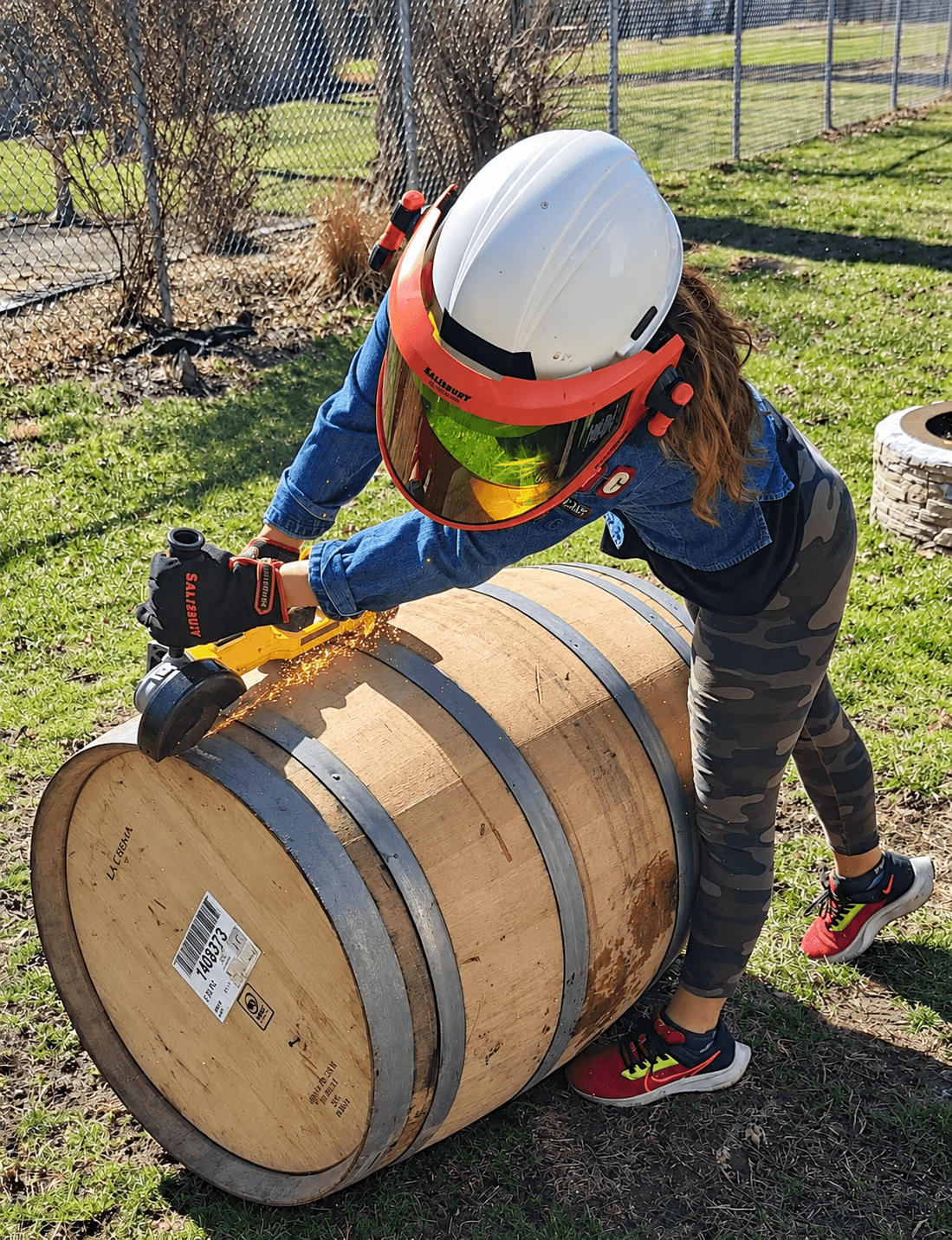 Child wearing a helmet and gloves rolling a large wooden barrel outdoors on grass.