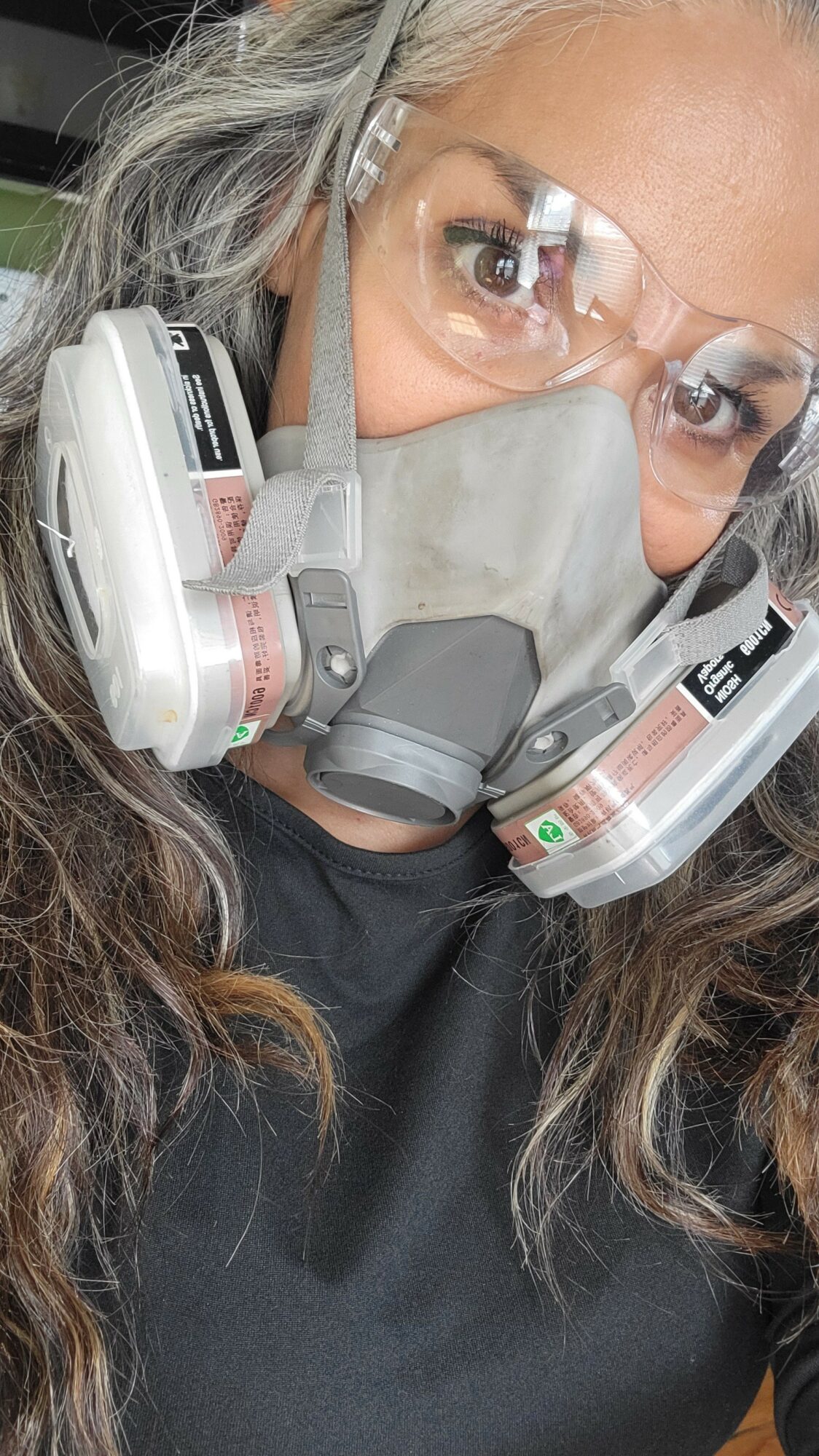 Person wearing safety goggles, respirator mask, and dual cartridge respirator, with gray hair and black clothing.