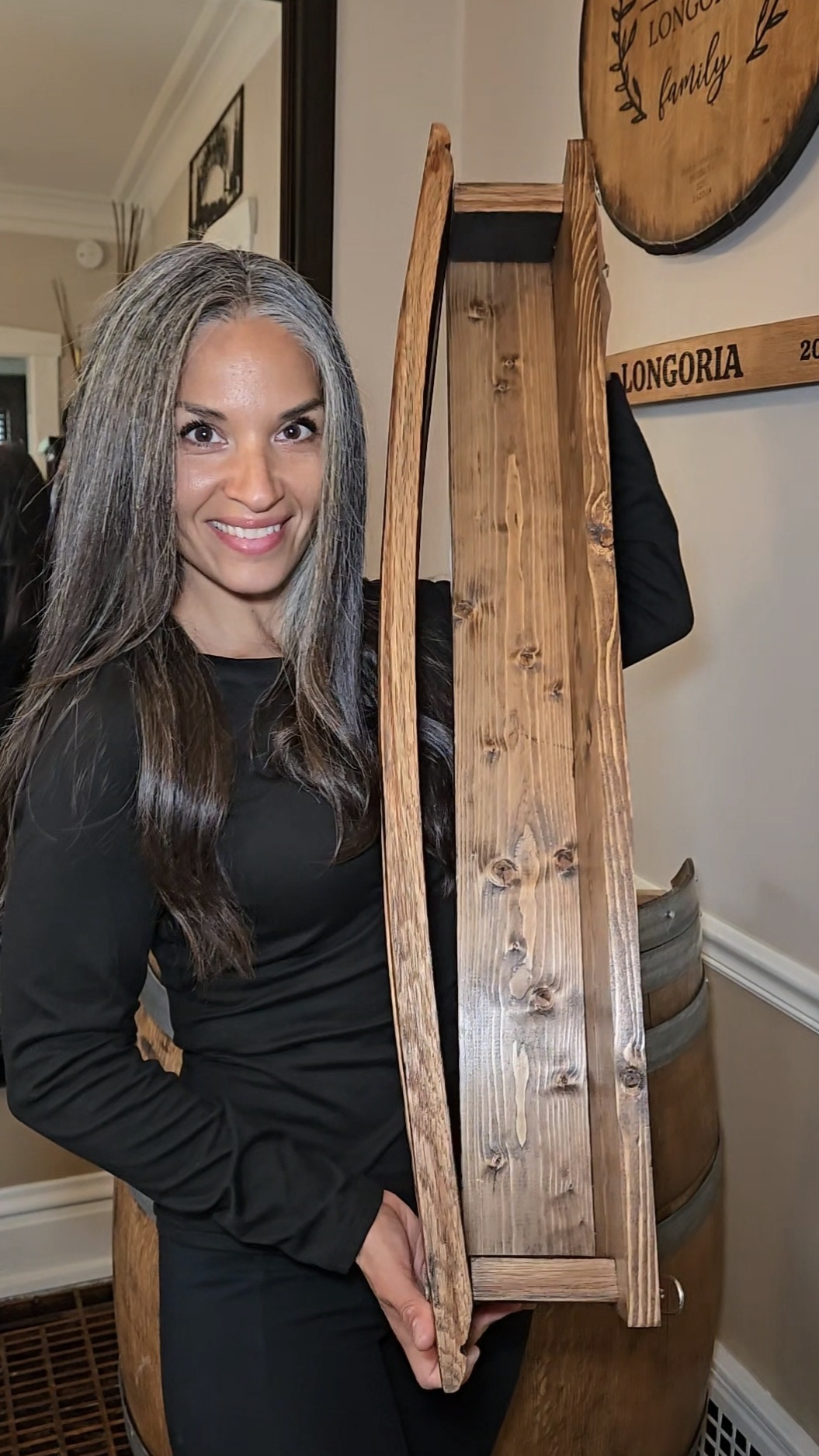 Woman with long gray hair smiling, holding a wooden boat-shaped object indoors.