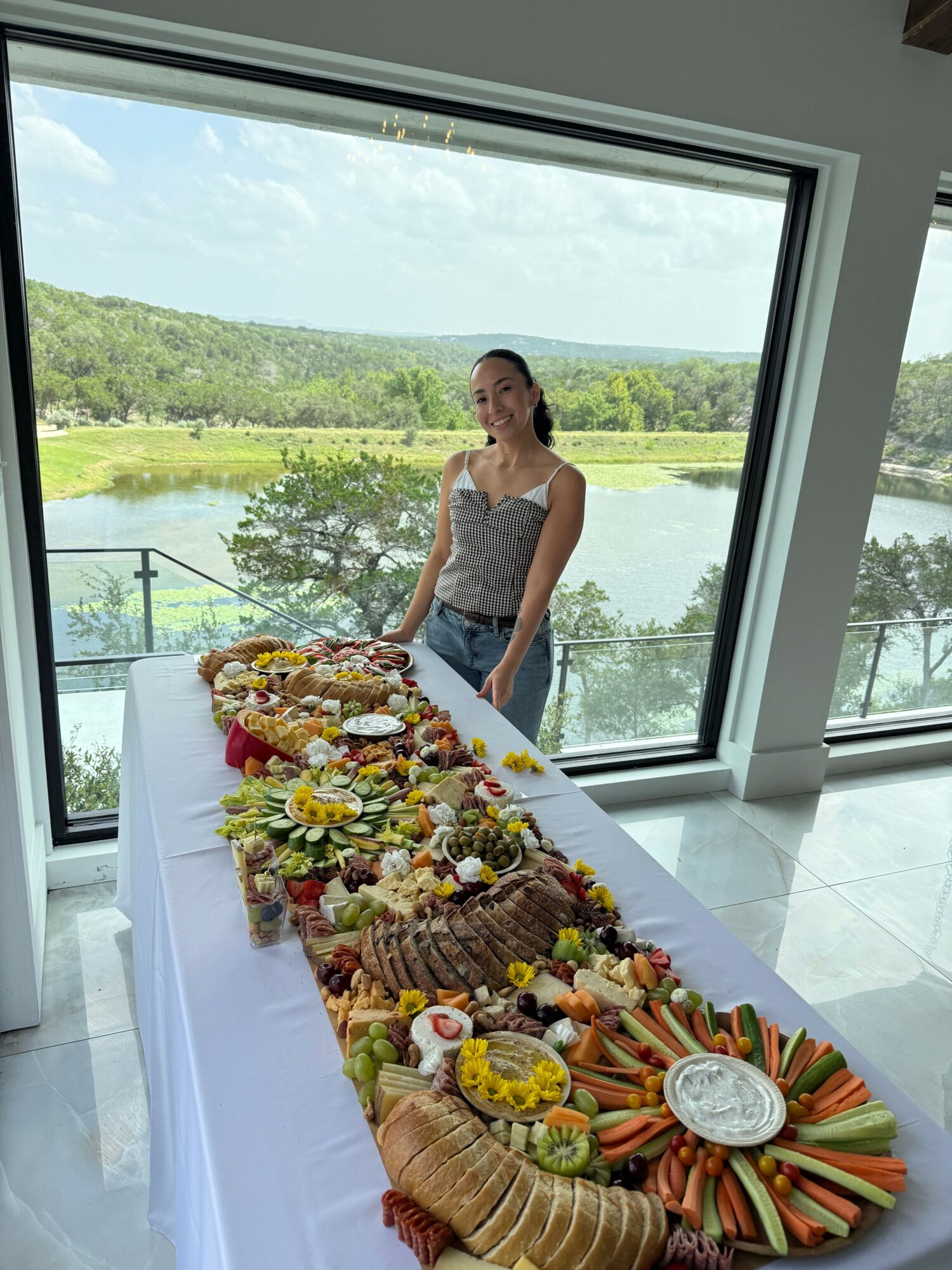 Woman standing near a table with a large decorated cake, overlooking a landscape through a window.