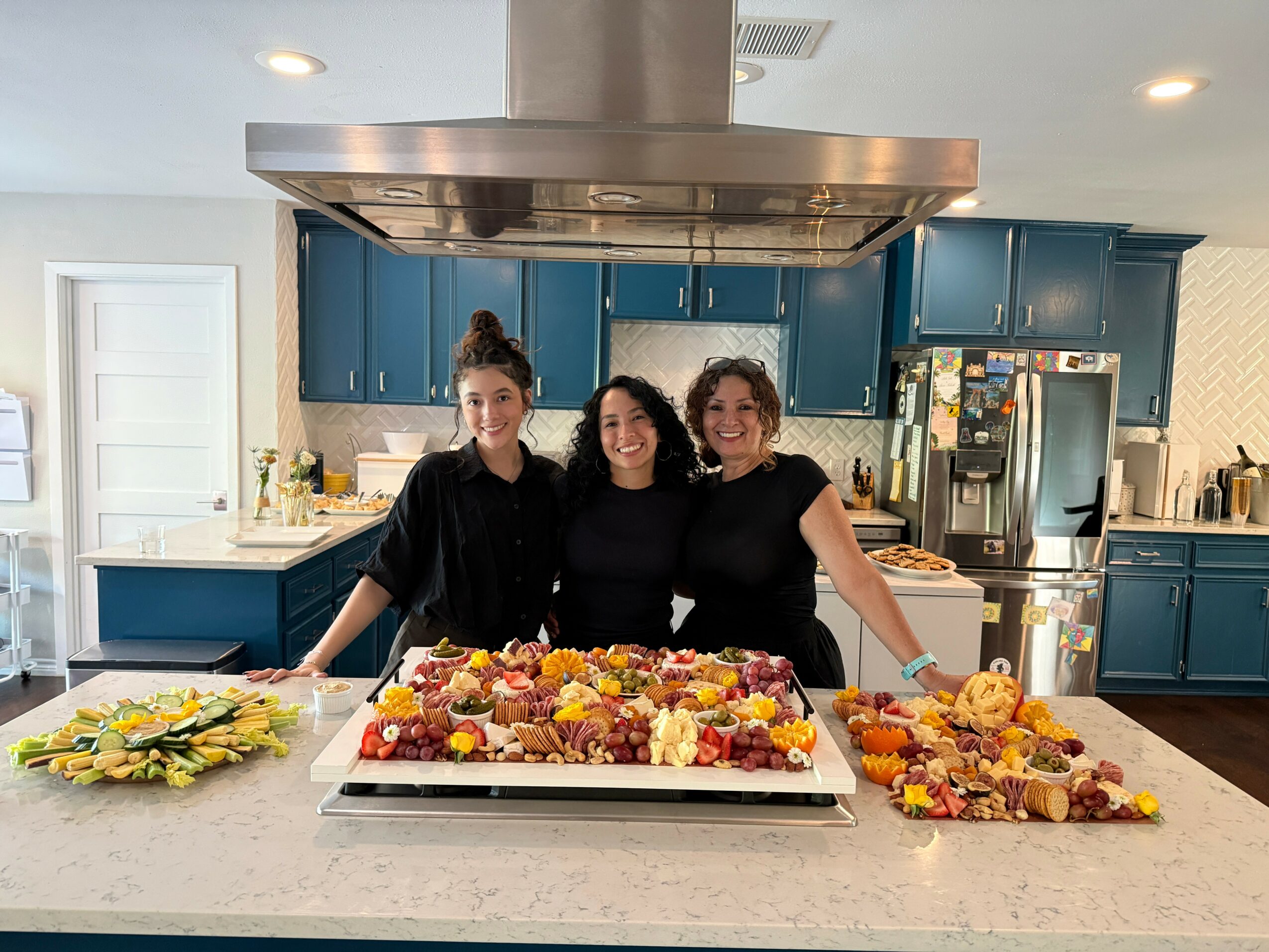 Three women standing behind a large decorated cake in a modern kitchen, smiling at the camera.