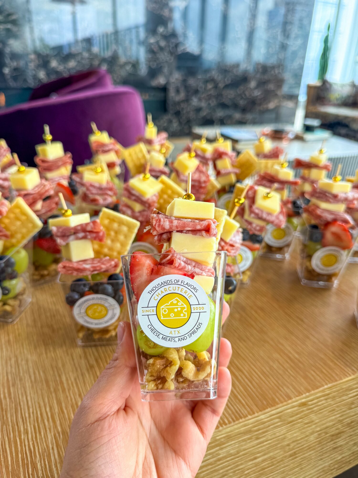 Hand holding a small clear container with yellow snack, in front of a table filled with similar containers and snacks, window in background.