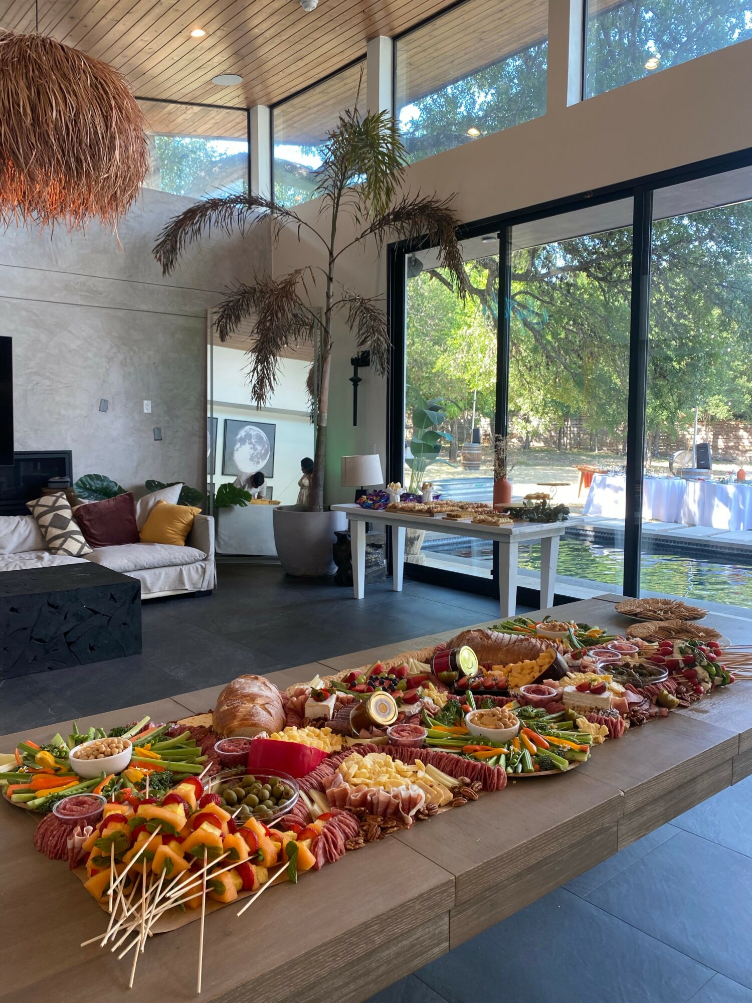 Long table with colorful food and skewers, large windows with trees outside, modern interior with plants and seating area.