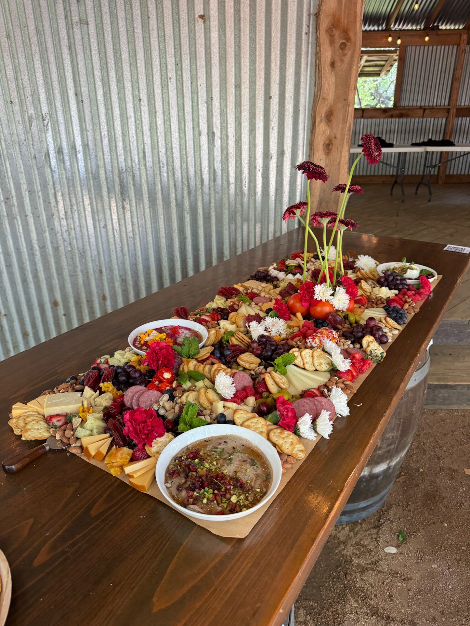 Colorful flower arrangement on a wooden table with bowls of sauce, set against a corrugated metal wall and rustic background.
