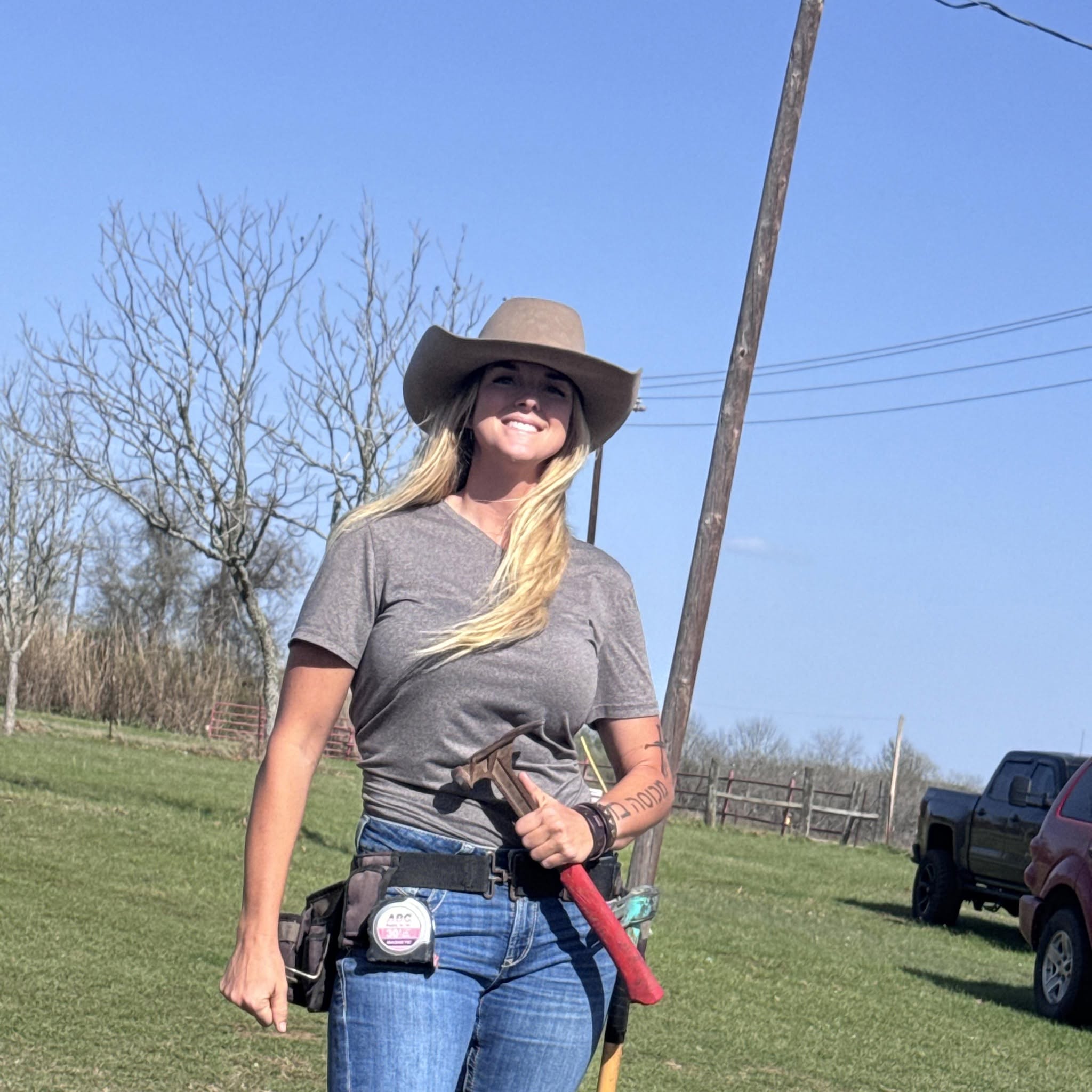 Woman wearing a wide-brimmed hat and gray t-shirt outdoors, holding a tool, with a clear blue sky and trees in background.