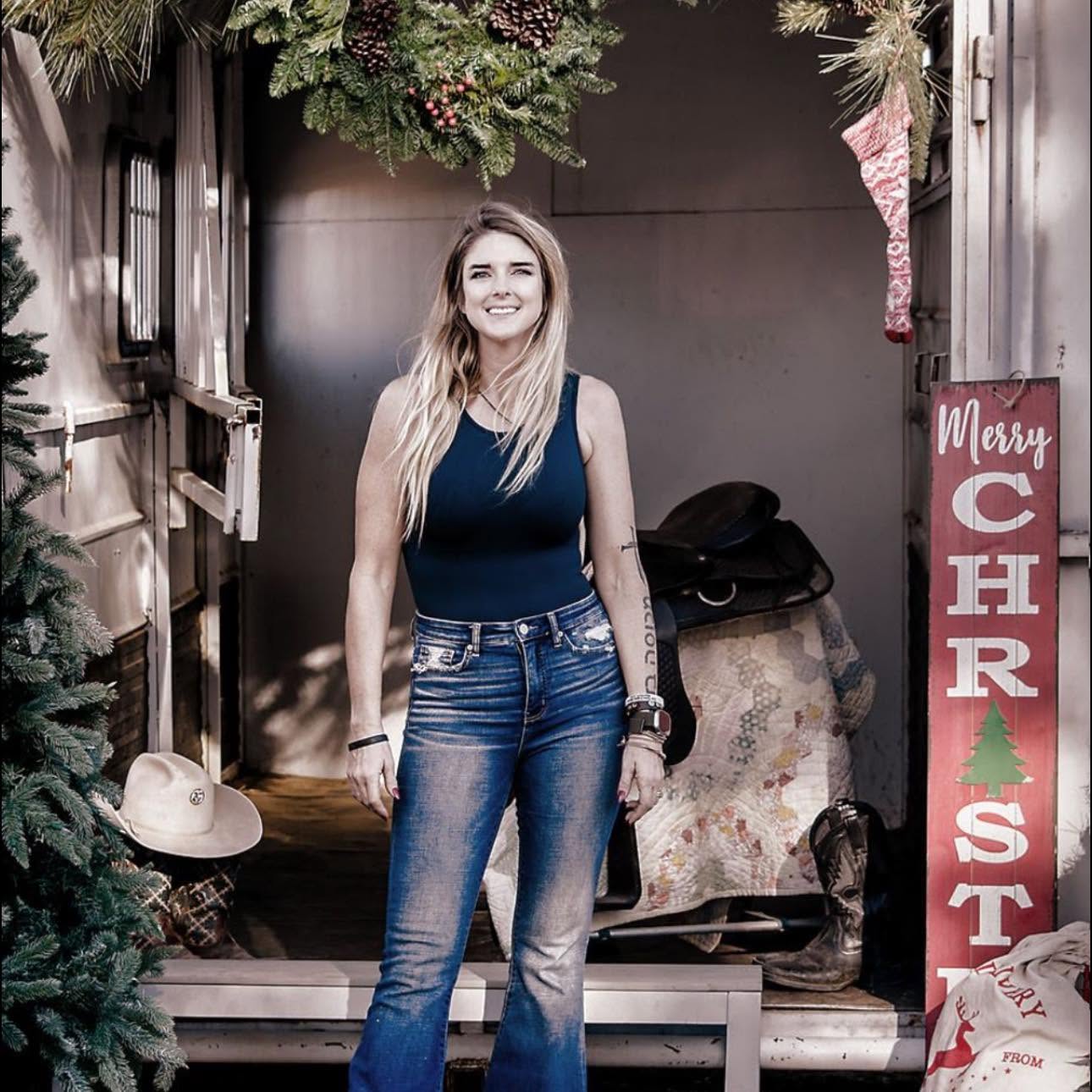 Young woman with long hair smiling, standing outdoors in front of a decorated background, wearing a sleeveless top and jeans.