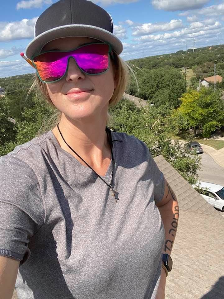 Woman wearing sunglasses and a cap outdoors, with trees and a parking lot in the background.