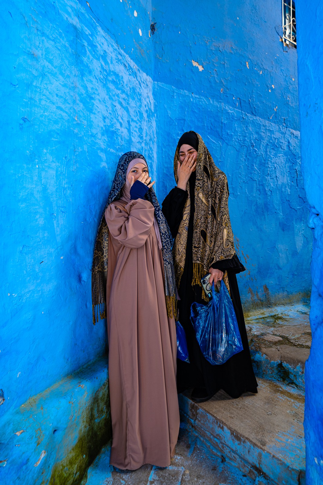 Two women stand against a blue wall, covering their faces with their hands, wearing long dresses and headscarves.