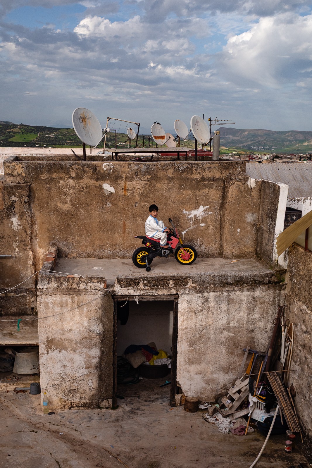 Child riding a small motorcycle on a rooftop with satellite dishes and a cloudy sky in background.