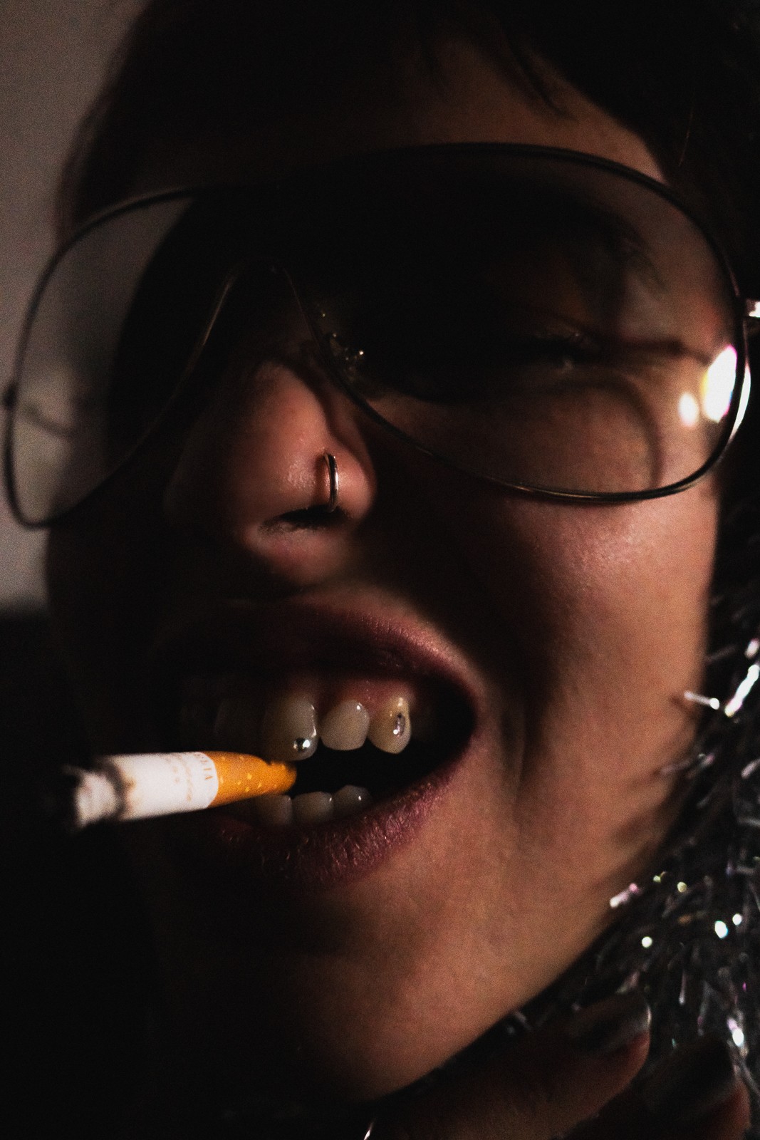 Person with glasses, nose ring, and cigarette in mouth, close-up of face with dark background.