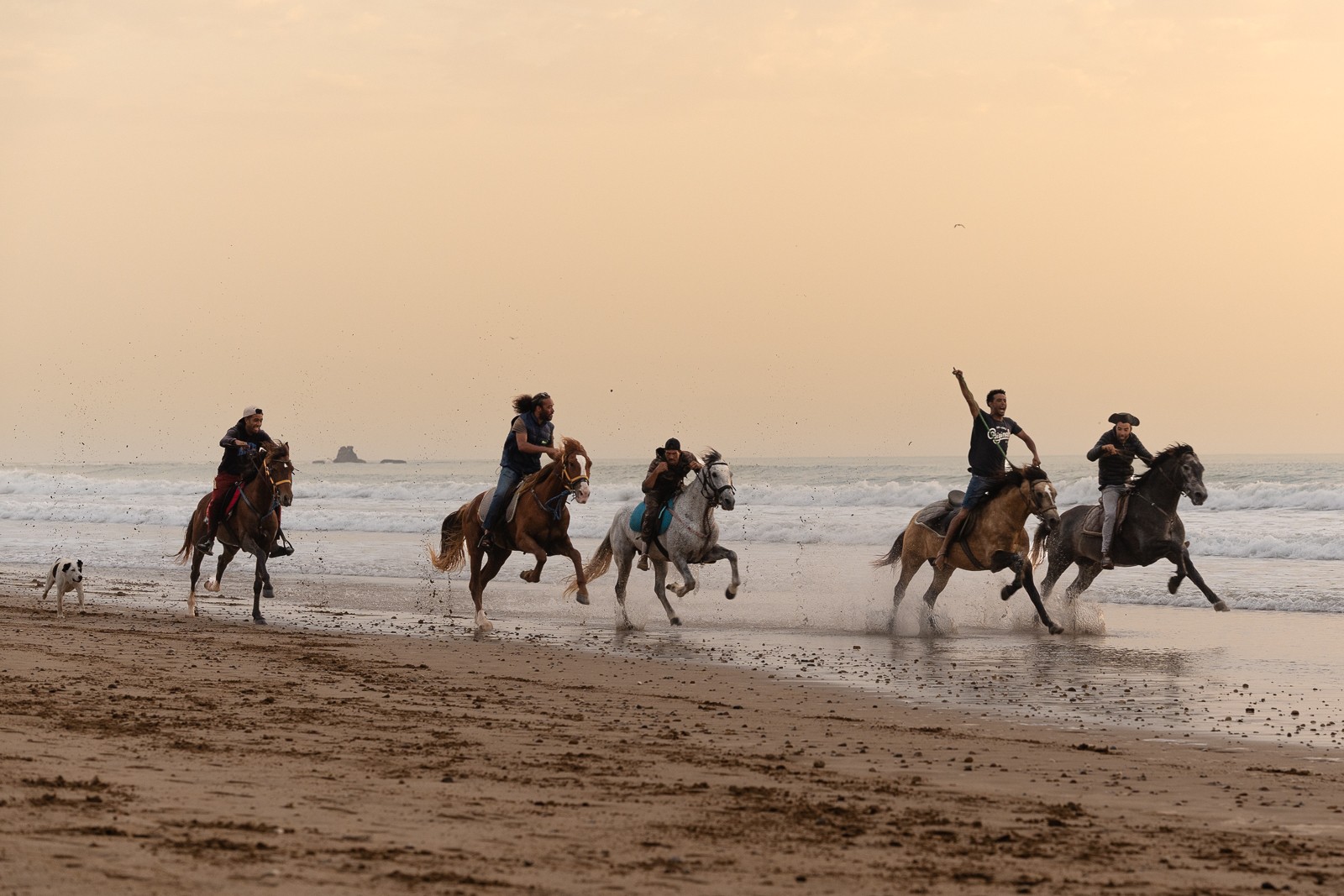 Group of people riding horses along the beach at sunset, some waving, with calm water and sky in background.