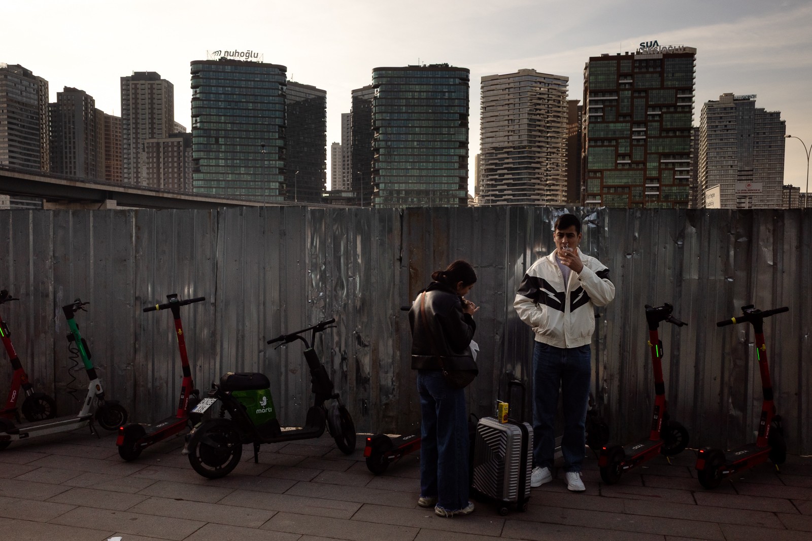 Two people standing near electric scooters and luggage against a city skyline with tall buildings, overcast sky.
