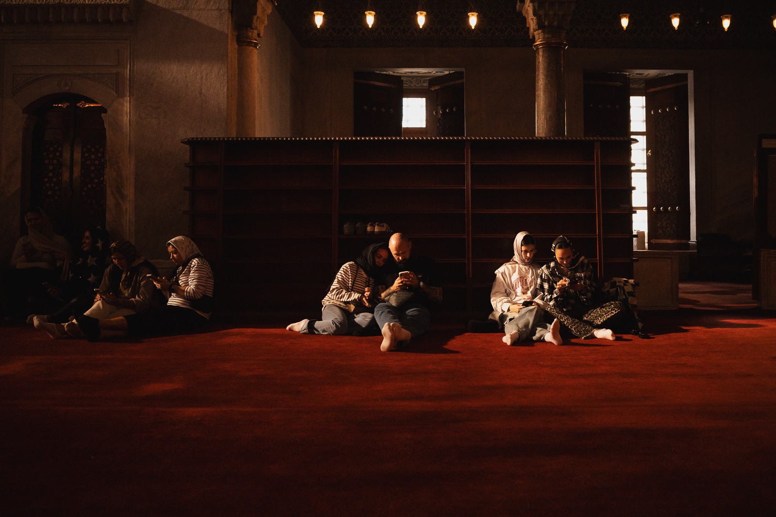 People sitting on the floor in a dimly lit room with tall windows and a large bookshelf behind them.