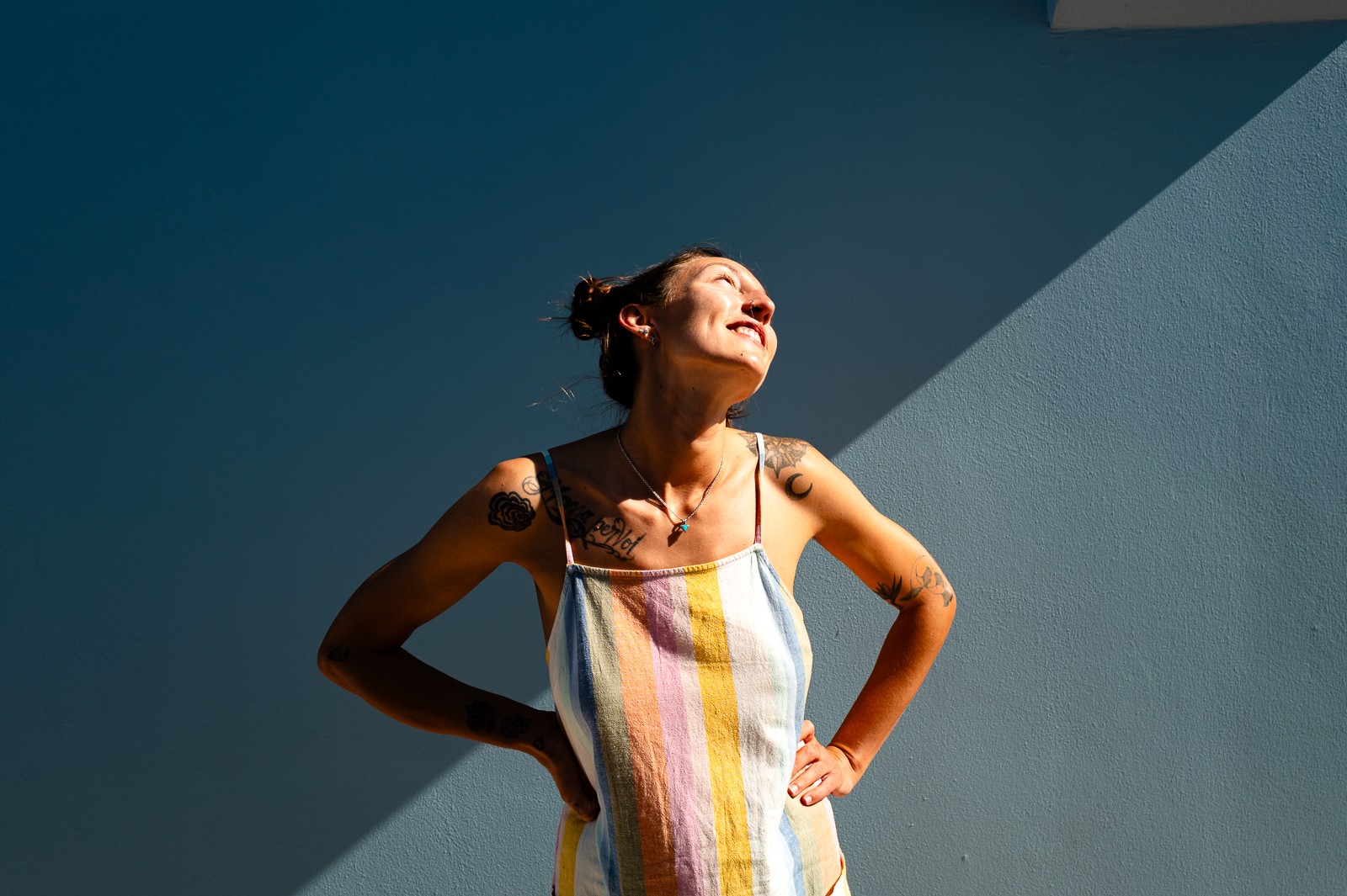 Woman standing outdoors with sunlight on her face, smiling, against a blue wall with a shadowed area.