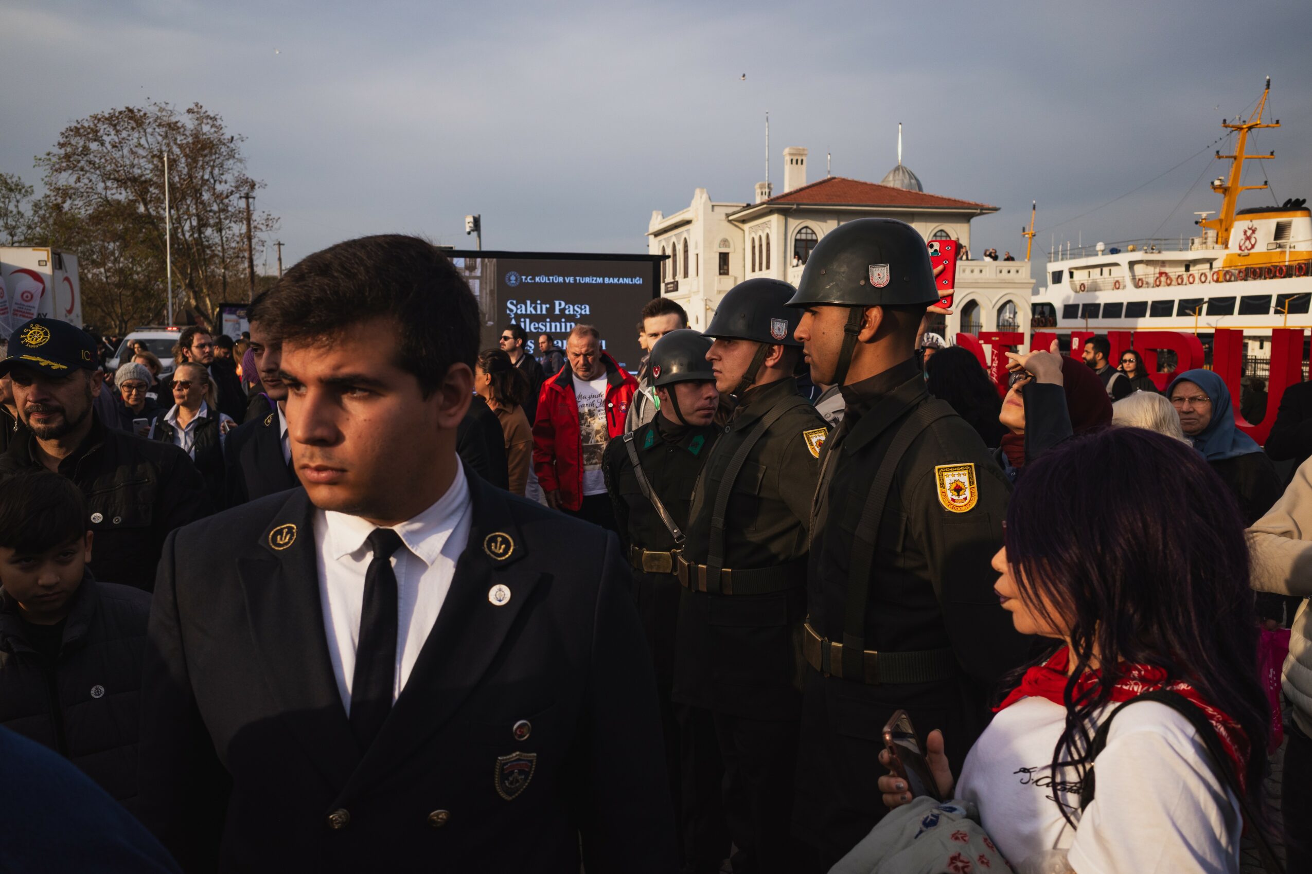 Group of uniformed officers and civilians outdoors near a ship, with a building and cloudy sky in background.