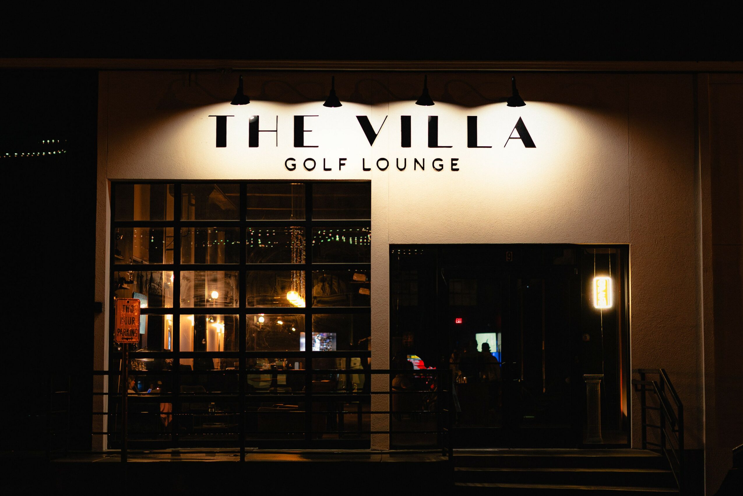 Sign for The Villa Golf Lounge illuminated at night, with a window showing interior lighting and reflections.