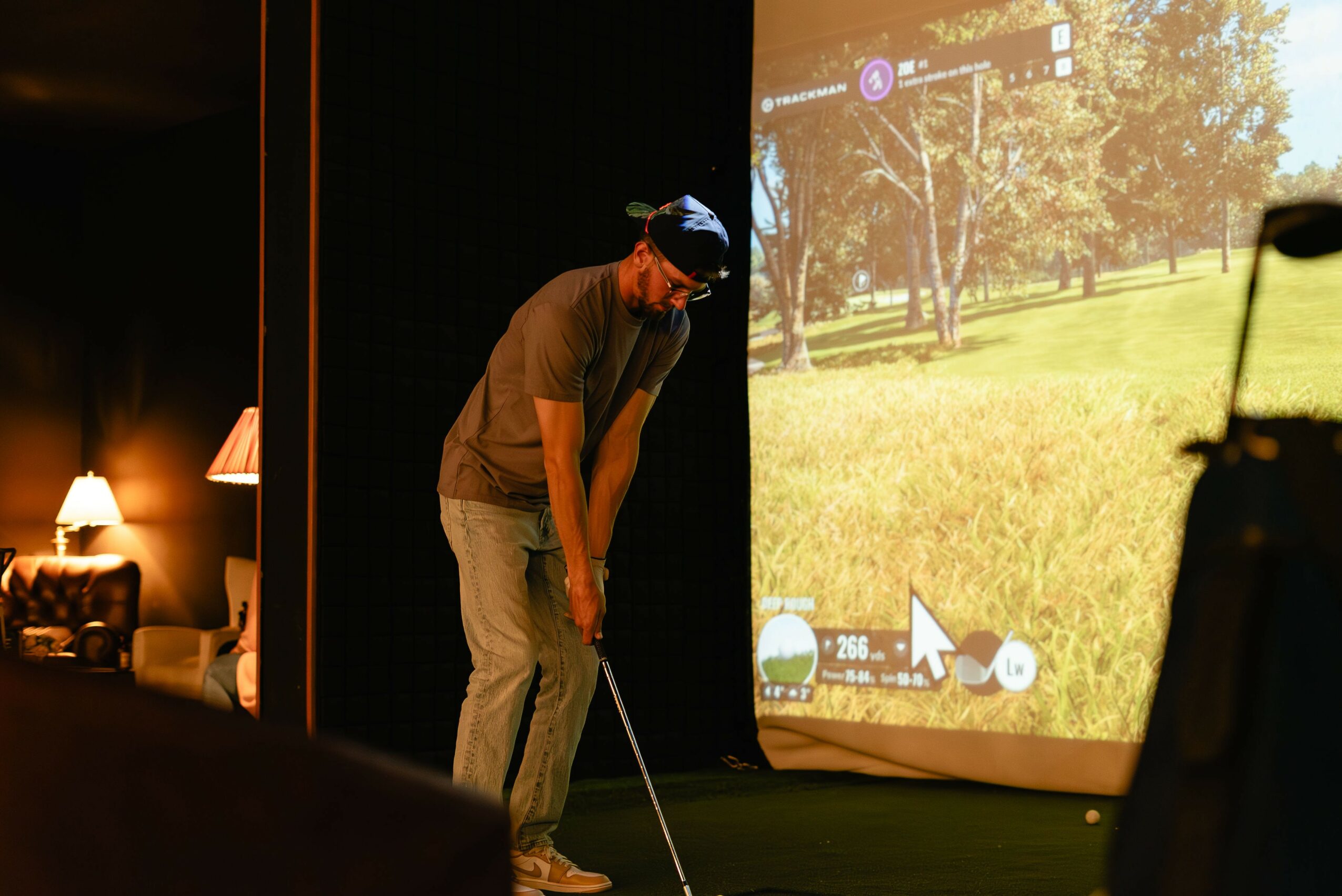 Person in a golf simulator room preparing to hit a golf ball, with a large screen showing a golf course.