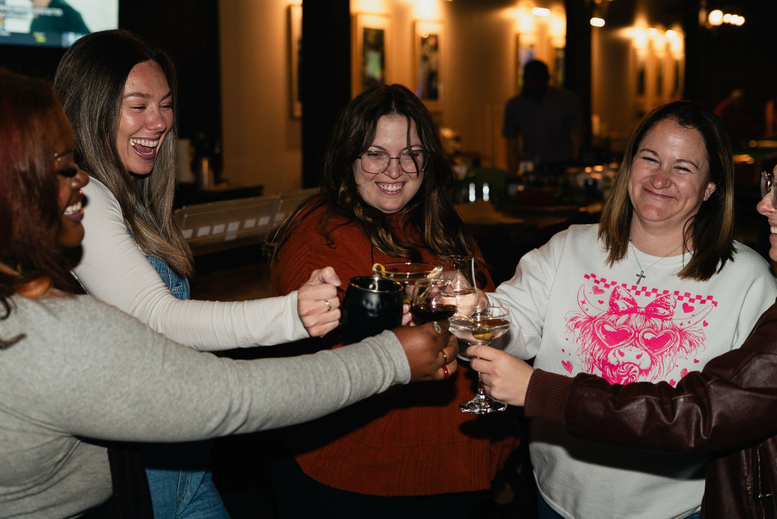 Four women smiling and clinking glasses together at a social gathering, warm lighting in the background.