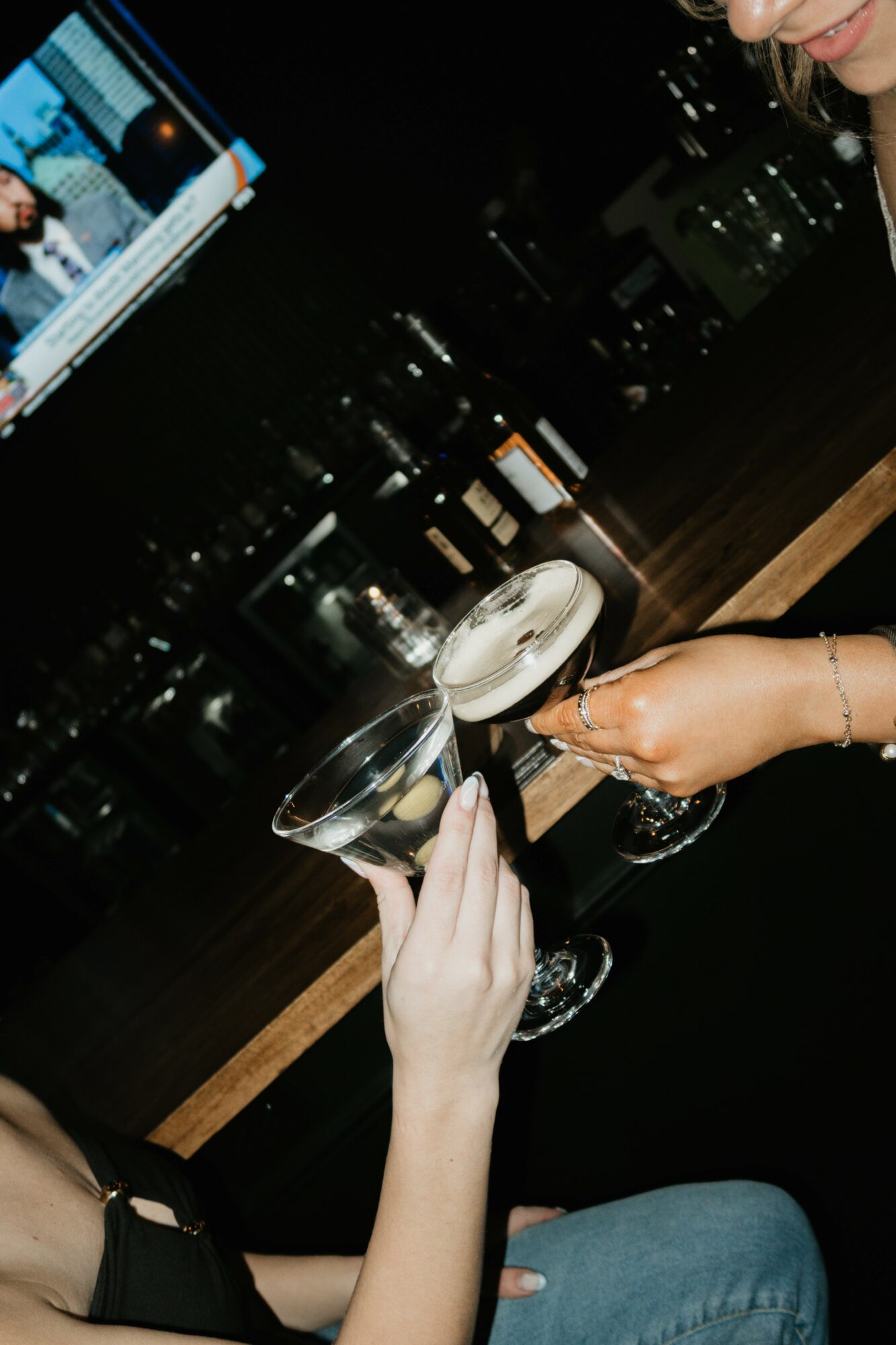 Two people clink cocktail glasses in a bar with city lights in the background.