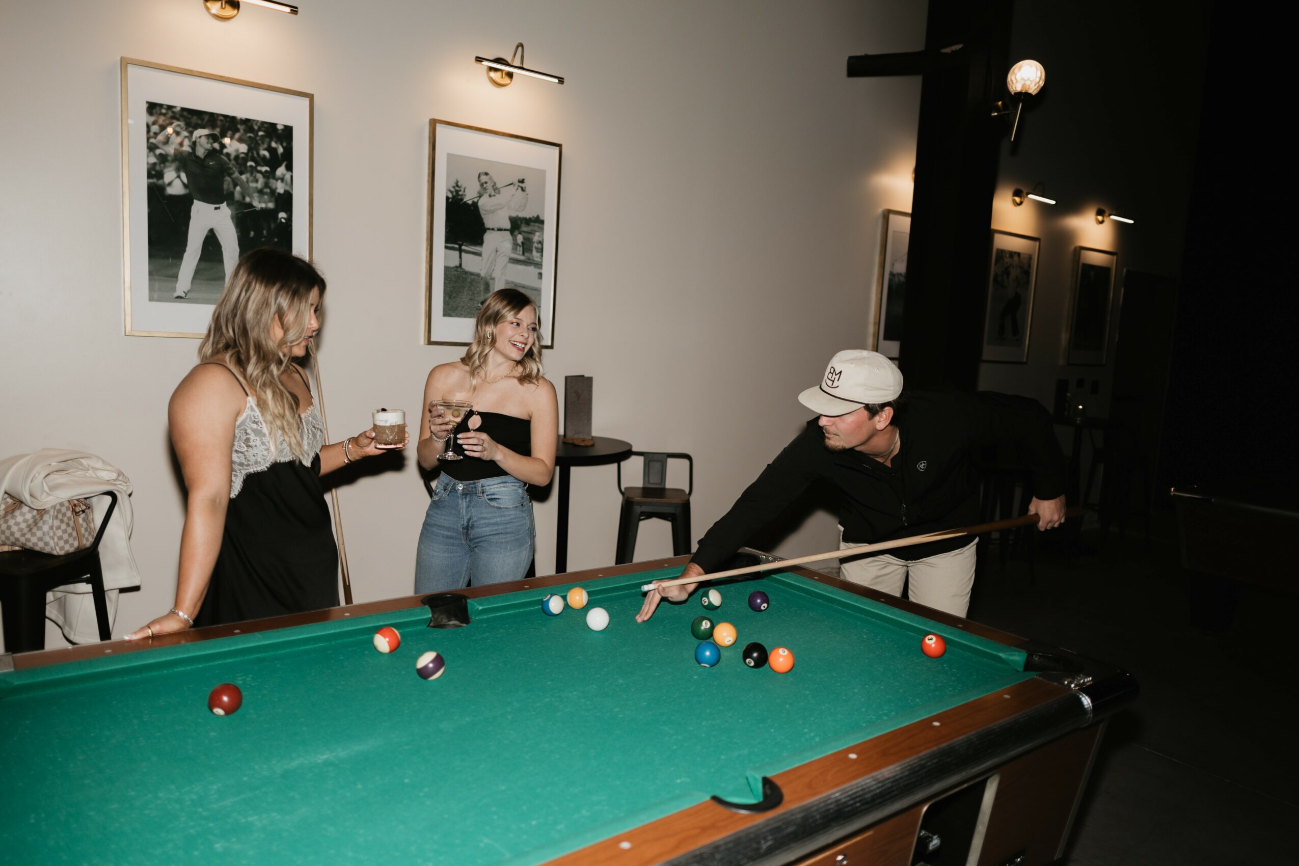 Three people around a pool table in a room with framed pictures on the wall, one person leaning over the table.