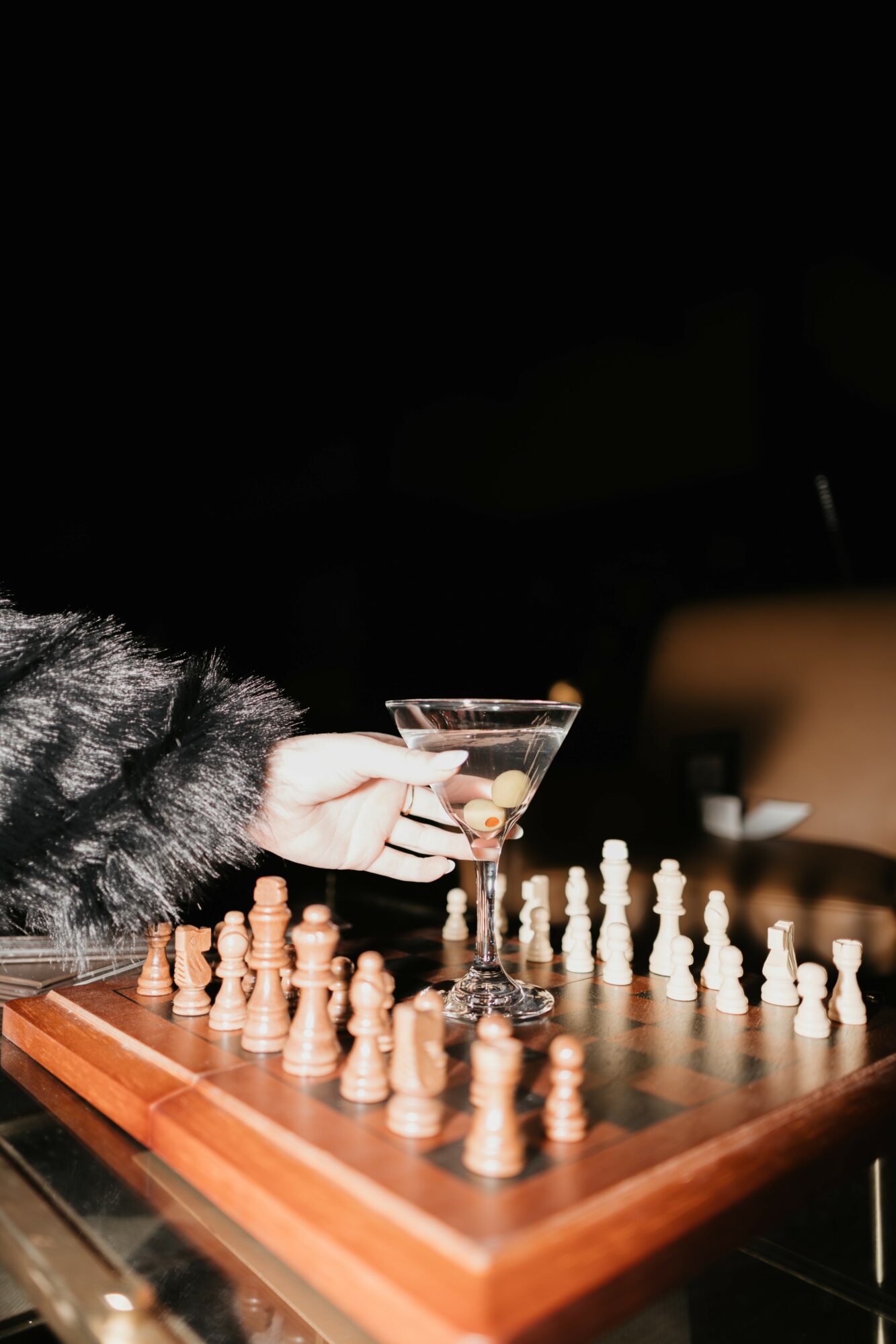 Person holding a martini glass over a chessboard with chess pieces, wearing a furry coat, in a dark setting.