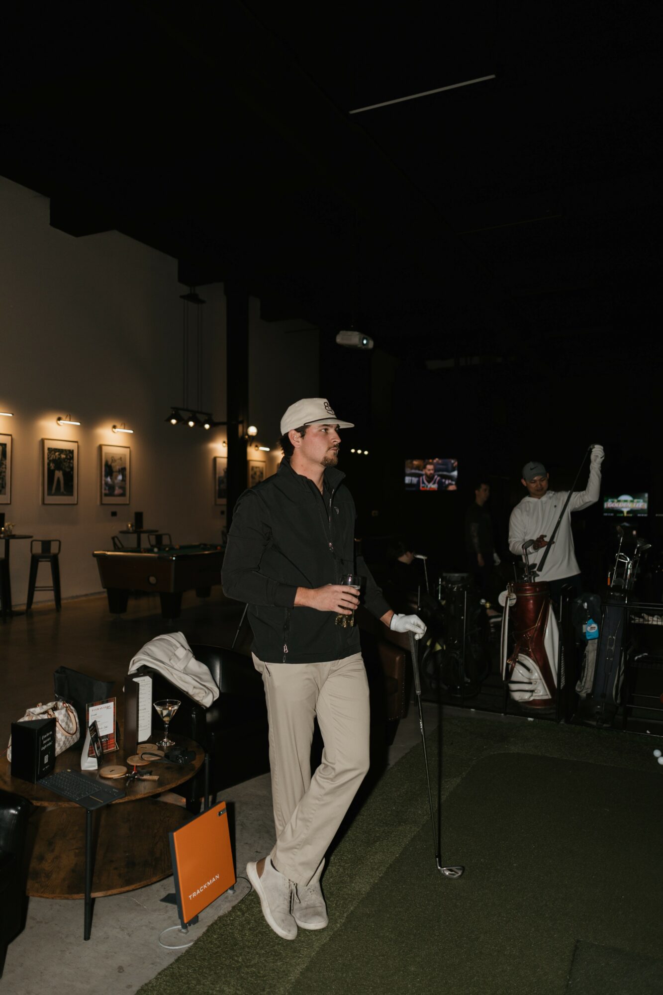 Woman in a hat and black jacket holding a drink, standing near a microphone in a dimly lit indoor space.