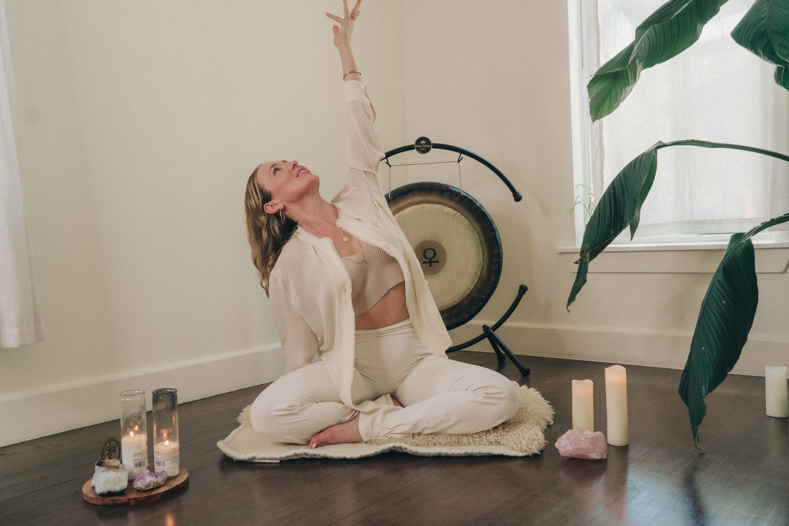 Woman practicing yoga indoors, sitting cross-legged on a mat, reaching upward with one arm, surrounded by candles and plants.