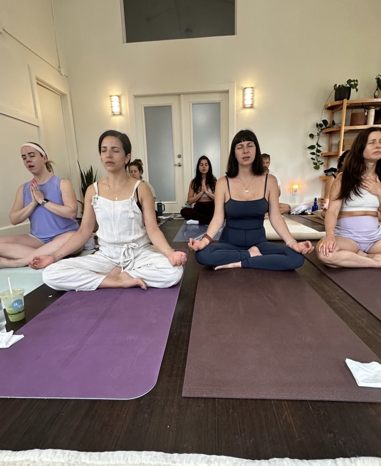 People practicing yoga in a bright room, sitting cross-legged on mats with hands in prayer position, eyes closed.