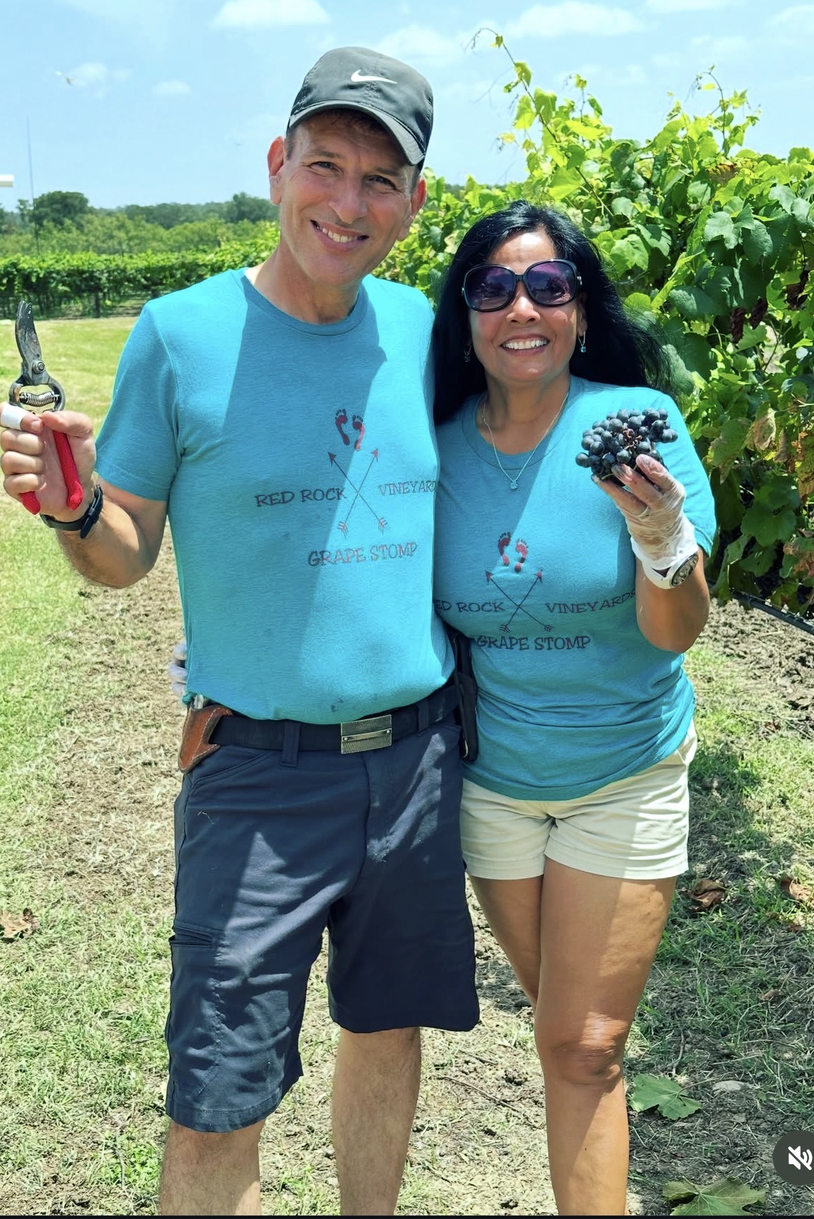 Two people outdoors, one holding a bunch of grapes, smiling, with green plants and blue sky in background.