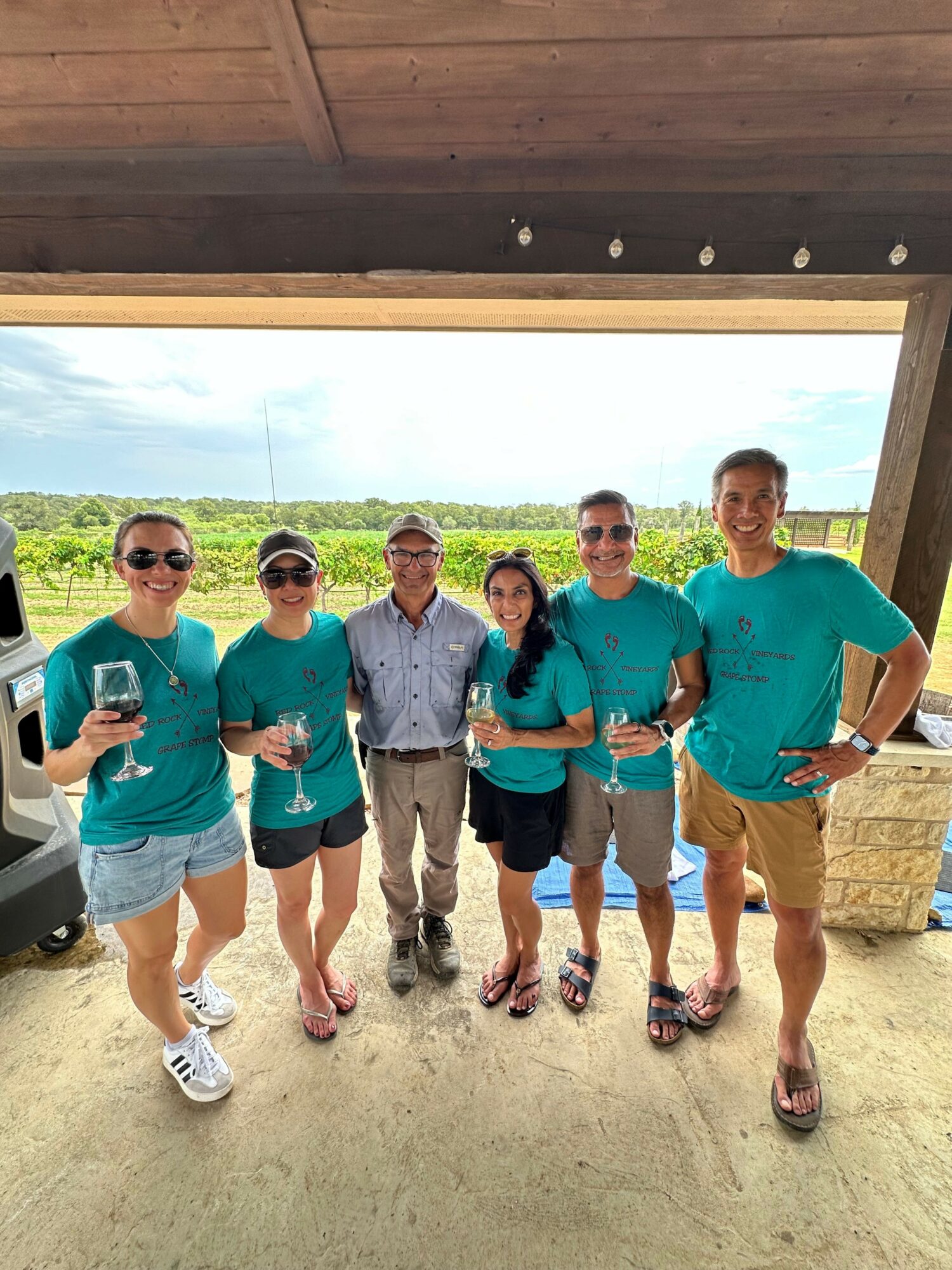 Group of seven people standing outdoors on a sandy surface, with a wooden structure and greenery in the background.